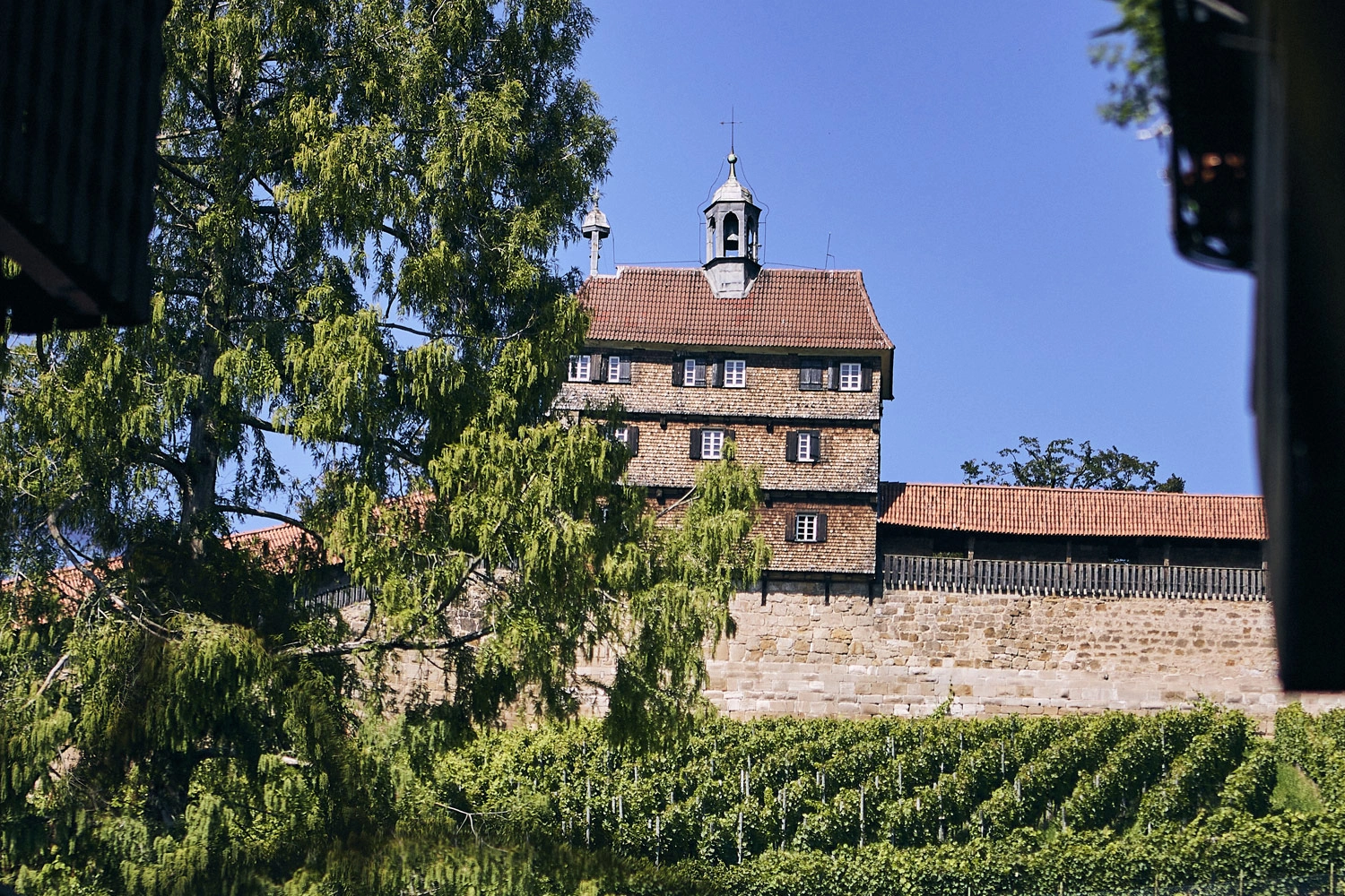 Historische Esslinger Burg mit Hochwacht, Fachwerkhaus und Stadtmauer über Weinreben, umgeben von Bäumen unter blauem Himmel.