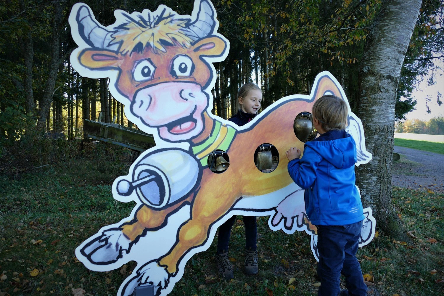 Zwei Kinder stehen vor einer Kuhfigur auf einem Wanderweg und läuten die daran angebrachten Glocken.
