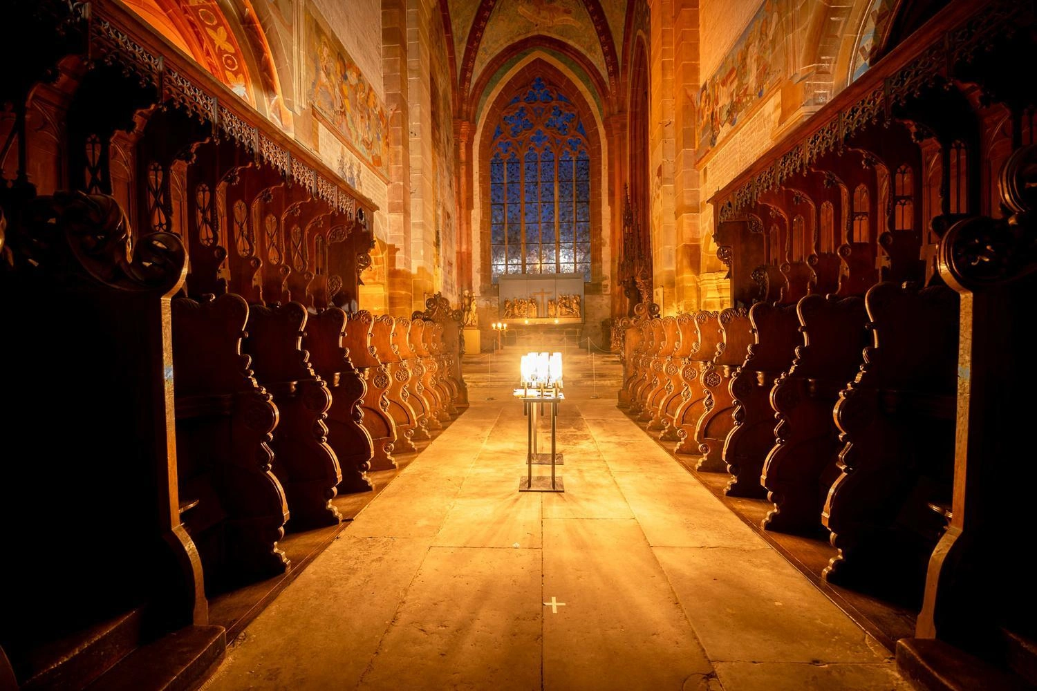 Beleuchtetes Kirchenschiff mit kunstvollen Holzschnitzereien, Kerzen im Mittelgang und farbigem Glasfenster im Hintergrund.