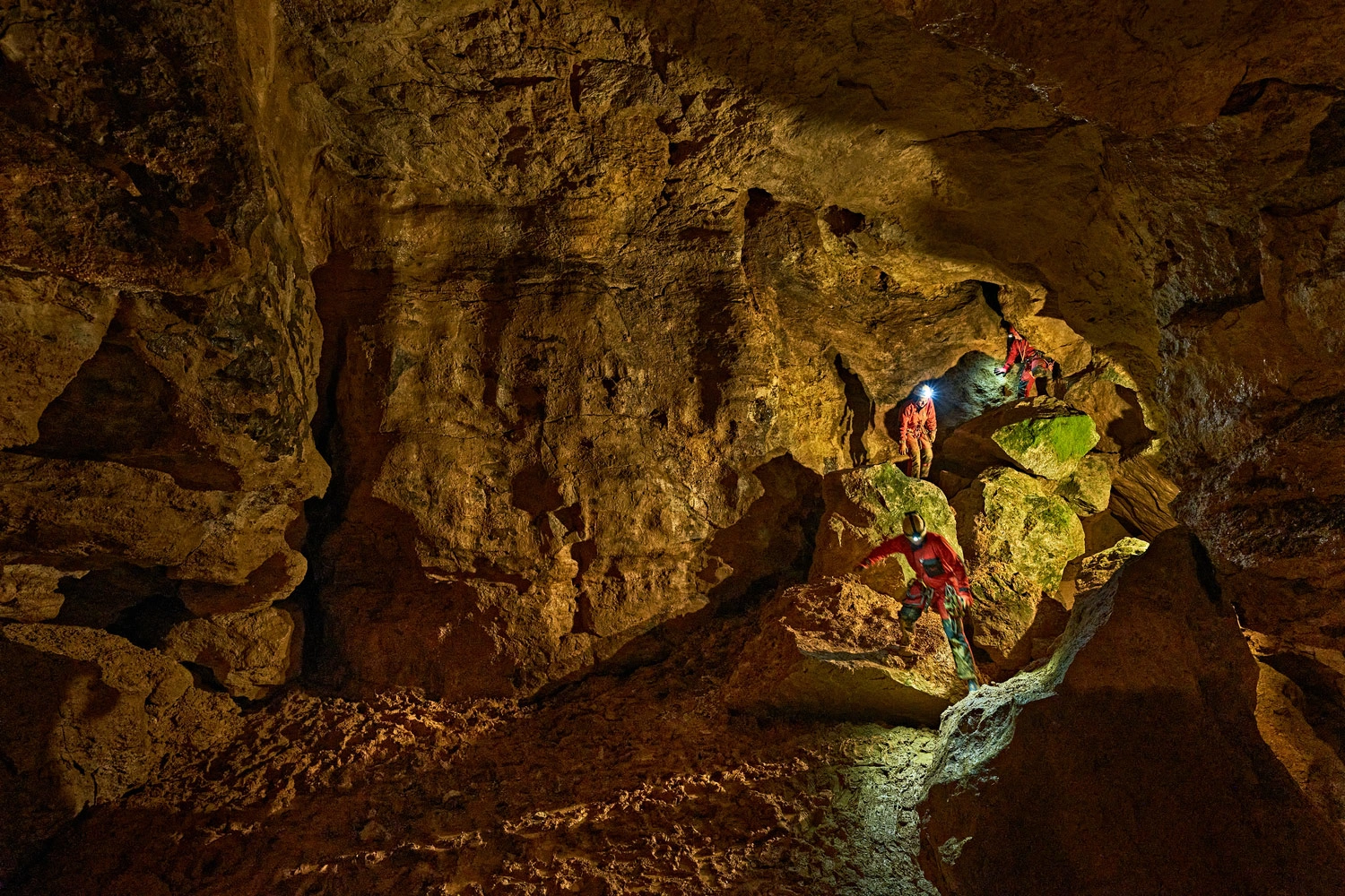 Gruppe von Personen in roten Overalls klettert mit Stirnlampen über große Felsen in einer weitläufigen, beleuchteten Höhle der Schwäbischen Alb.