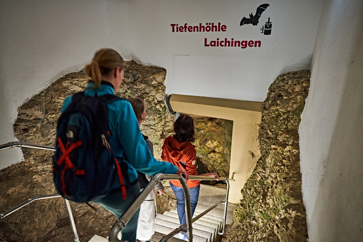 Zwei Personen steigen über eine Treppe mit Metallgeländer in die Tiefenhöhle Laichingen hinab, durch einen Eingang im Fels mit Beschriftung an der Wand.
