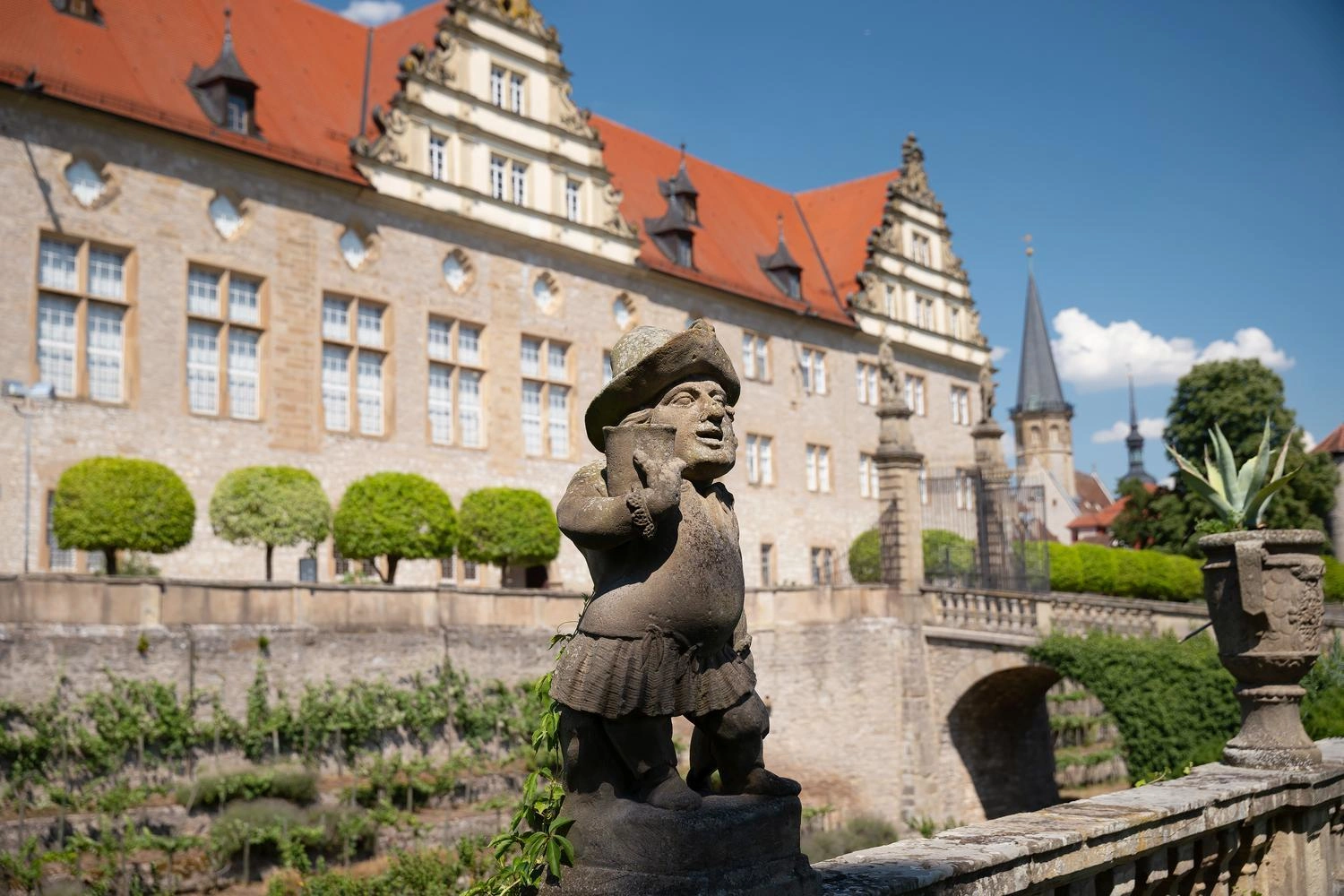 Steinfigur im Zwergengarten vor dem barocken Schloss Weikersheim mit rotem Dach, Brücke und gepflegten Gartenanlagen unter blauem Himmel.