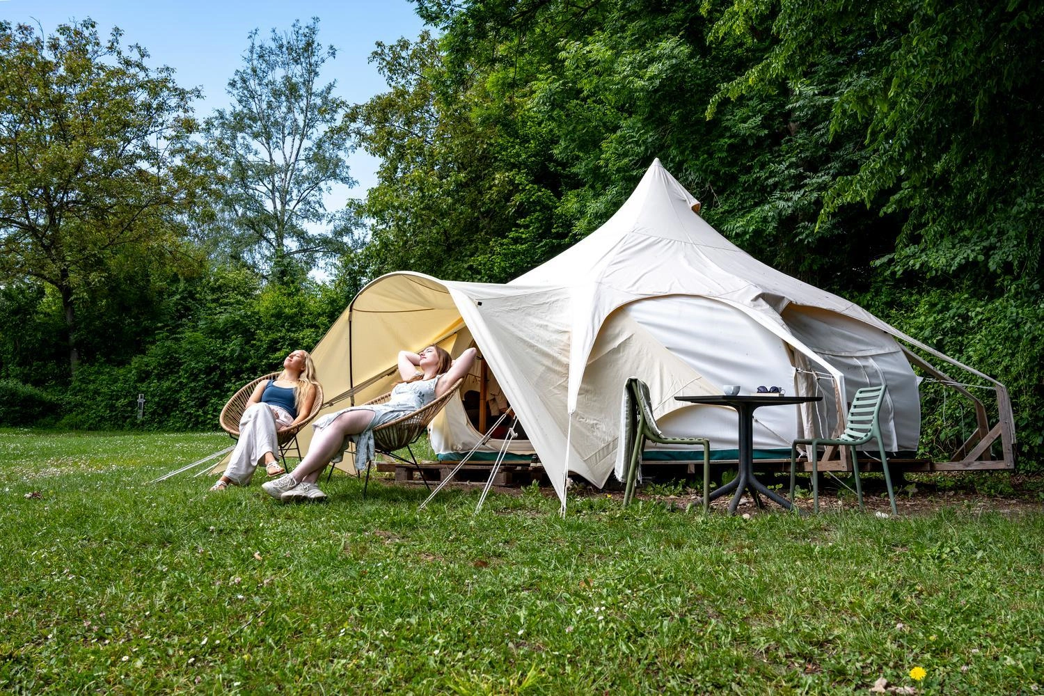 Zwei Personen entspannen vor einem großen cremefarbenen Glamping-Zelt auf einer Wiese am Waldrand, daneben stehen Gartenstühle und ein Tisch.