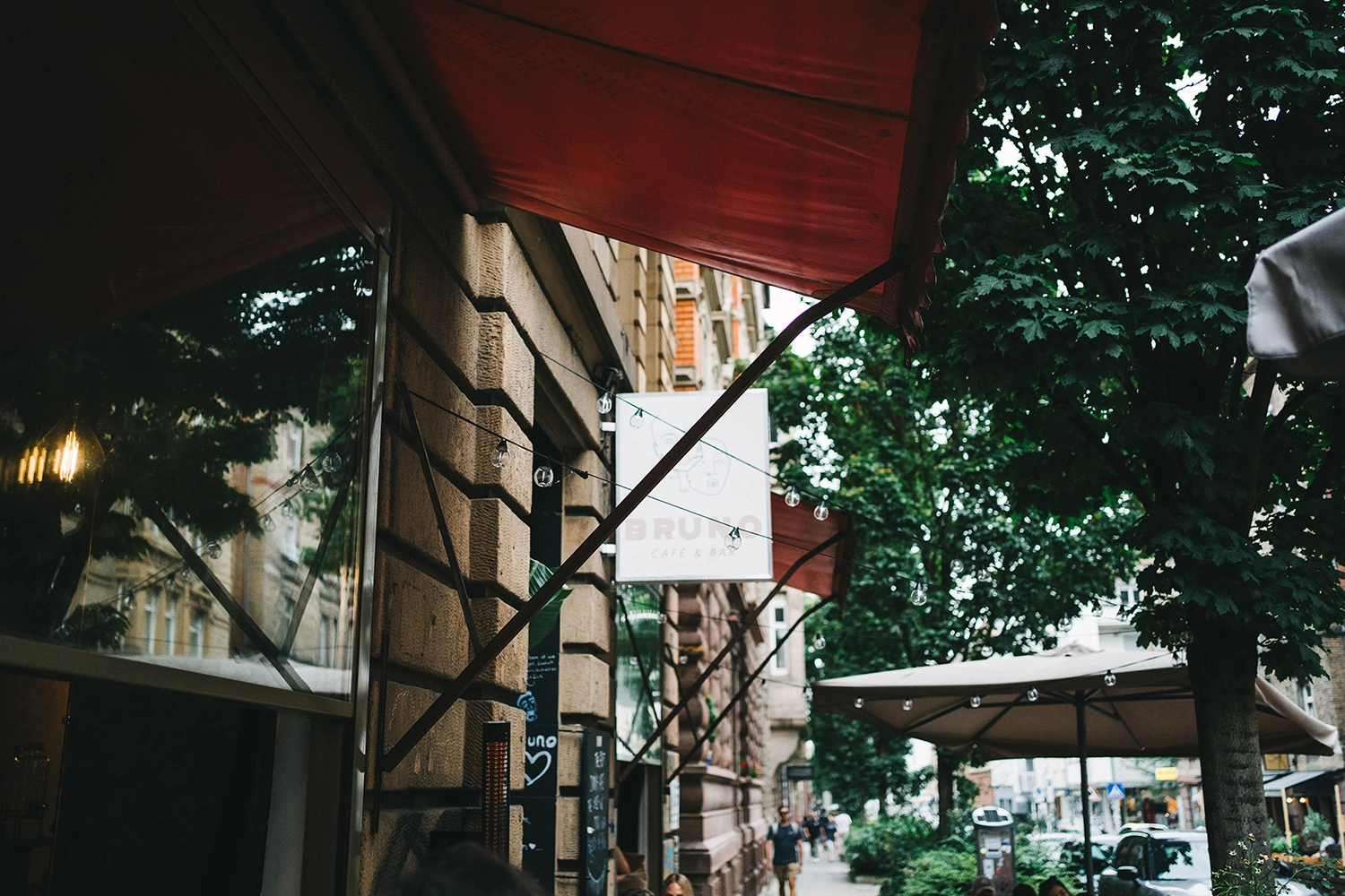 Rote Markise eines Straßencafés unter dichtem Laub, mit Lichterkette und Sitzplätzen im Außenbereich.