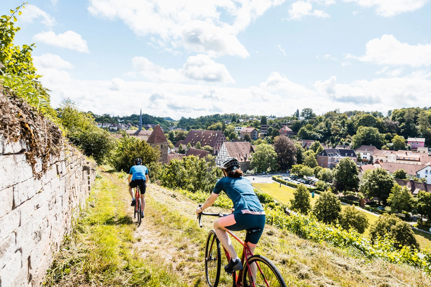 Zwei Gravelbiker fahren durch Weinberge. Im Hintergrund ist eine Klosteranlage zu sehen.
