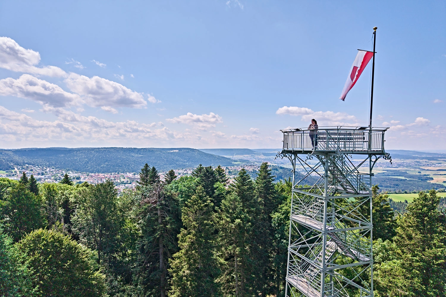 Person steht auf der Aussichtsplattform des Lembergturms mit wehender rot-weißer Fahne, Blick über Wälder und Hügel der Schwäbischen Alb unter blauem Himmel.