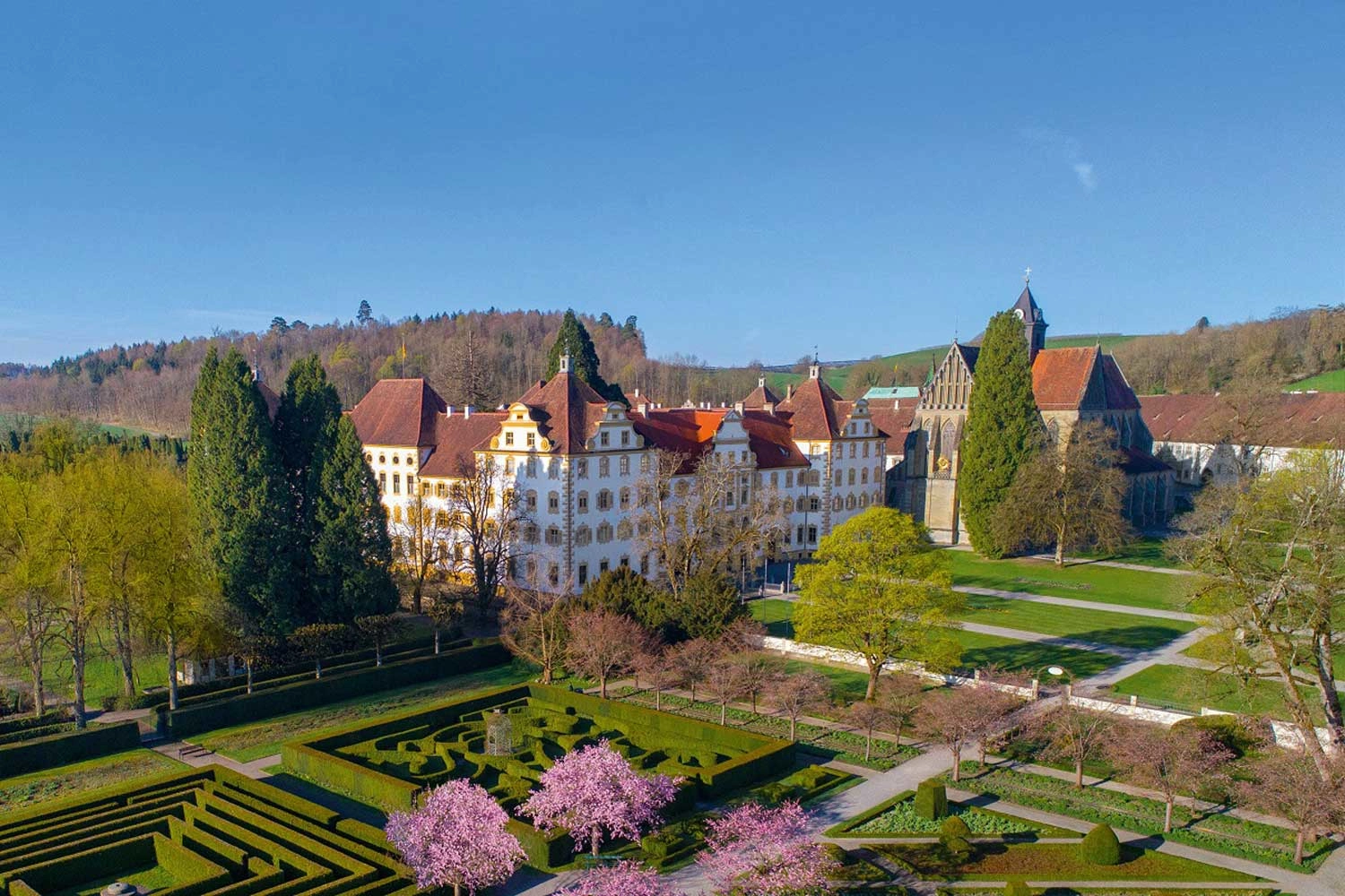 Luftaufnahme eines weißen Schlosses, davor ein akurater Graten mit rosa blühenden Bäumen.