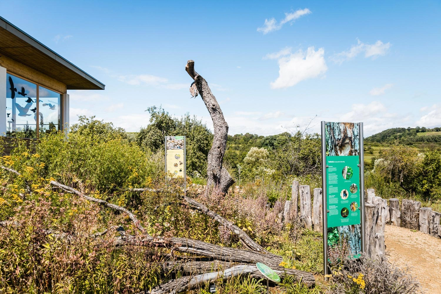 Eingang zu einem Naturparkzentrum. Auf dem Gelände gibt es wildes Gebüsch und abgebrochene Baumstämme.