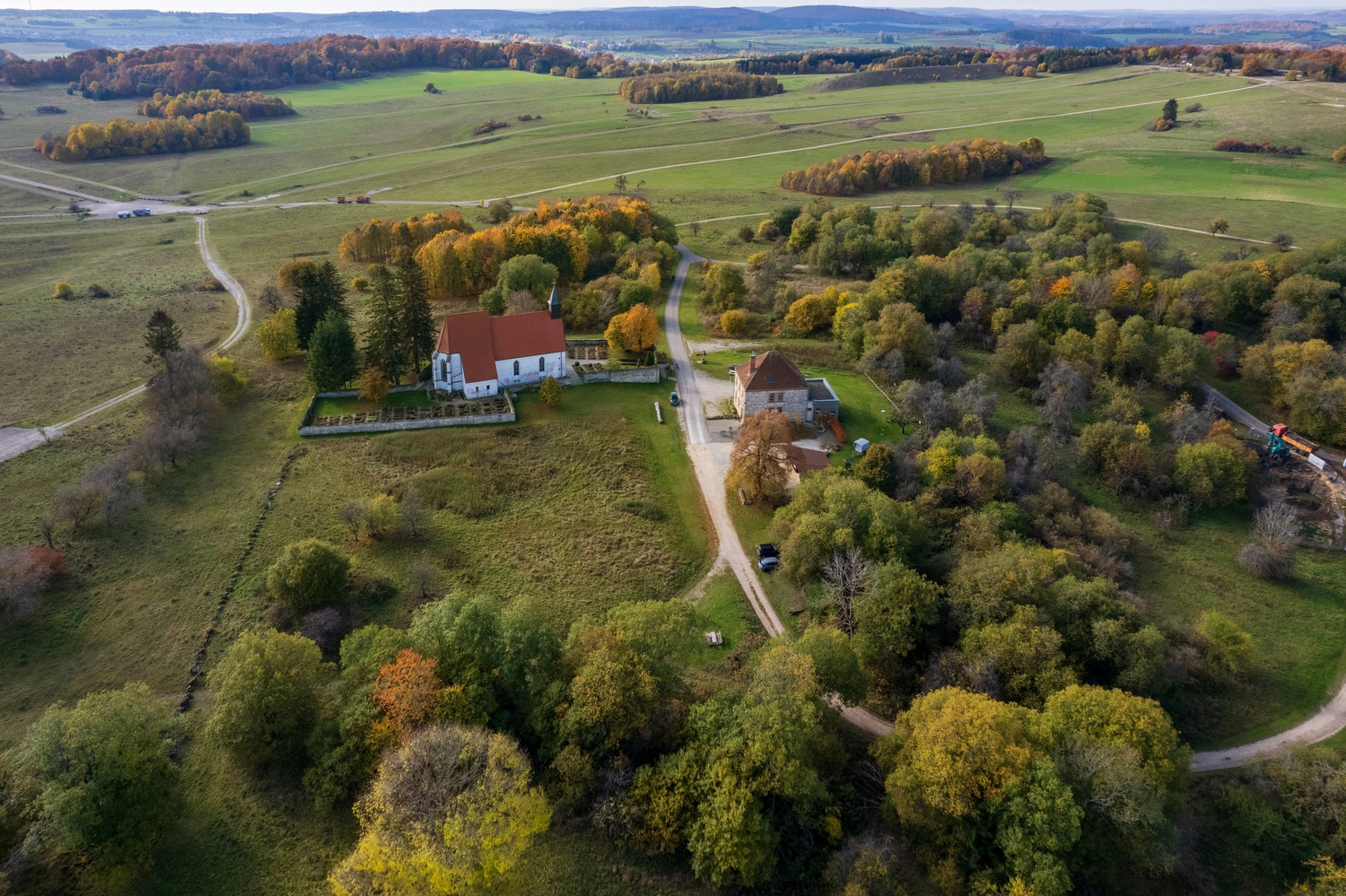 Luftaufnahme eines kleinen historischen Areals mit Kirche und Gebäuden, umgeben von herbstlichem Wald und weitläufigen Wiesen in sanfter Hügellandschaft.