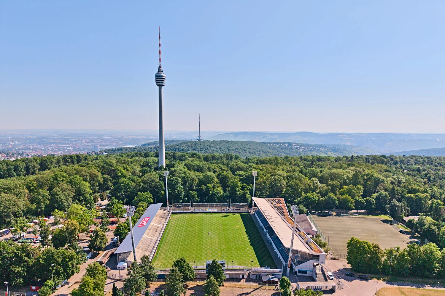 Luftaufnahme des Kickers-Stadions in Stuttgart, umgeben von Wald, mit dem Fernsehturm im Hintergrund unter klarem blauem Himmel.