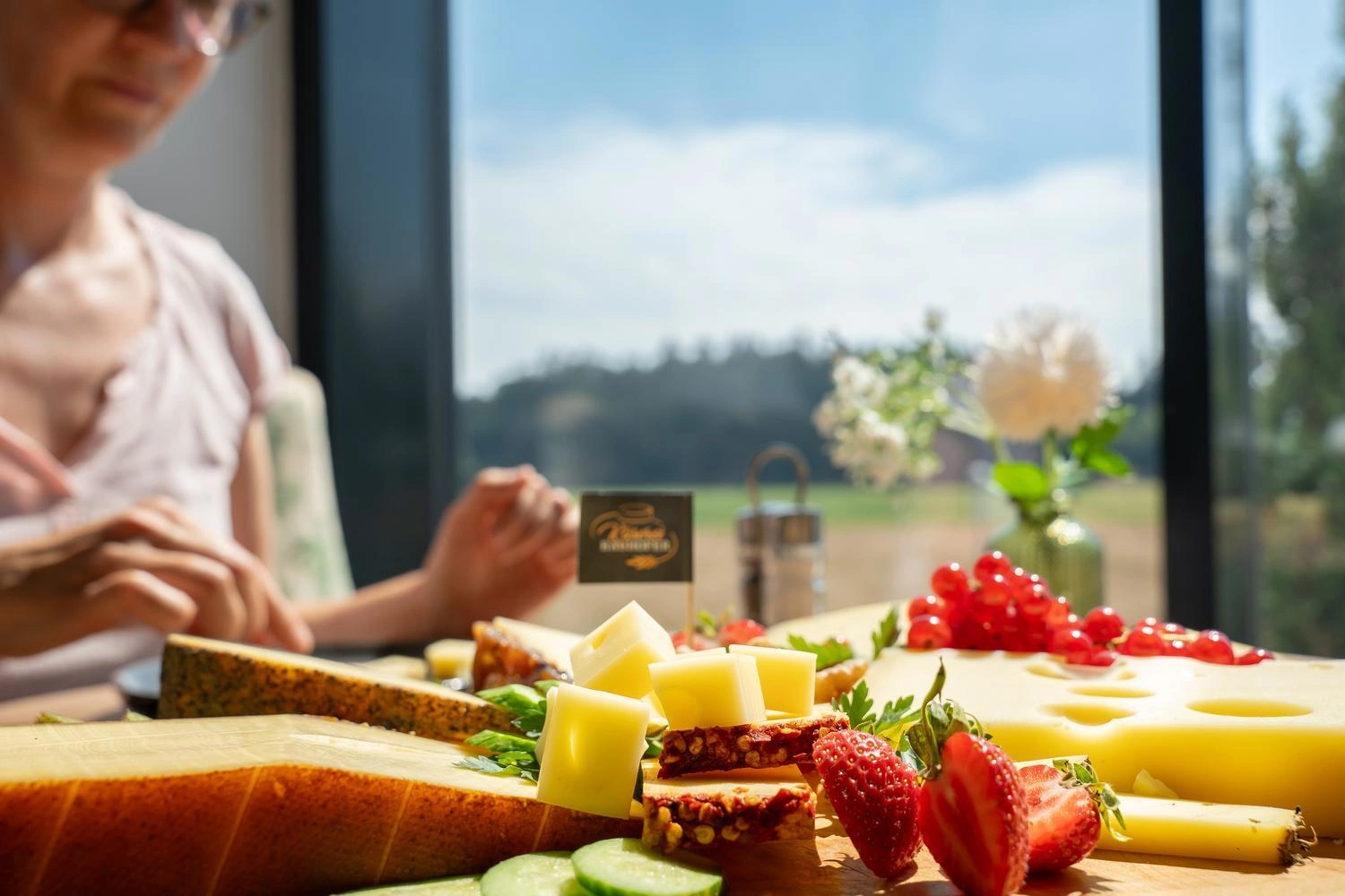 Frühstückstisch mit verschiedenen Käsesorten, frischen Erdbeeren, Gurkenscheiben und roten Johannisbeeren vor einem Fenster mit Blick ins Grüne.