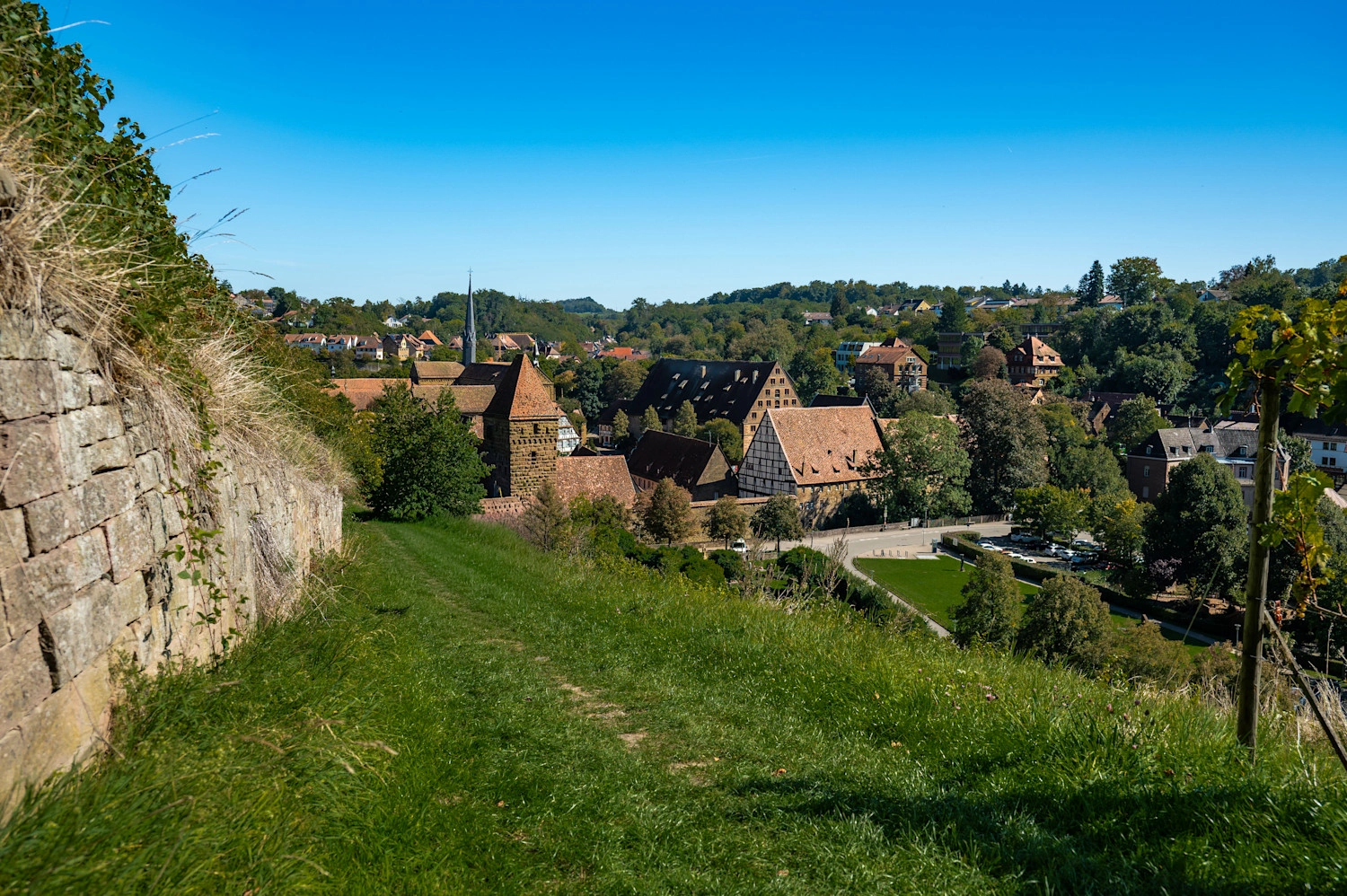 Wanderweg auf Rasen entlang einer steinernen Mauer. Im Hintergrund sind alte Gebäude zu sehen.