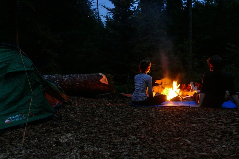 Two people are sitting by a campfire at a small campsite in the woods after dark.