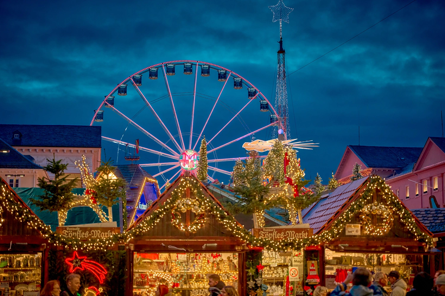 A row of illuminated stalls at a Christmas market. A Ferris wheel turns behind it.