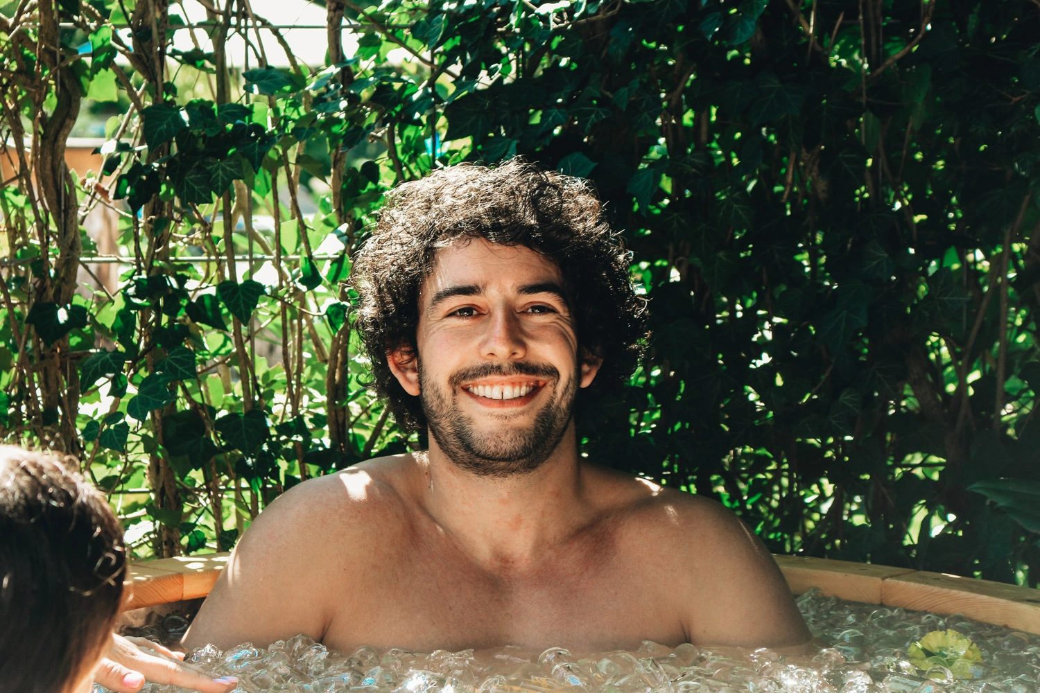 Person entspannt in einem sprudelnden Außen‑Whirlpool, umgeben von dichtem grünen Blattwerk und natürlicher Umgebung.