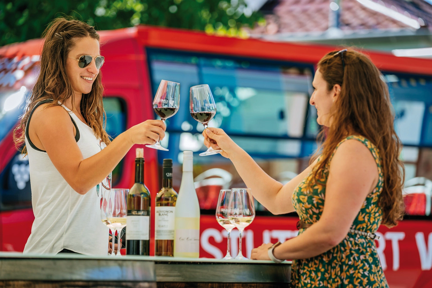 Zwei Frauen in sommerlicher Kleidung stehen an einem Bartisch und stoßen mit einem Glas Wein an.