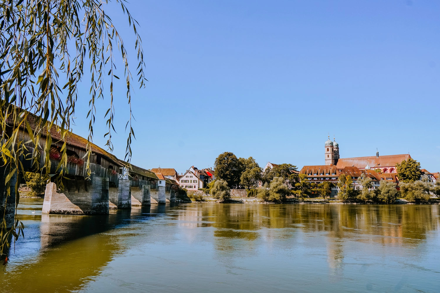 View across a river to an old town. On the left a covered wooden bridge crosses the river.