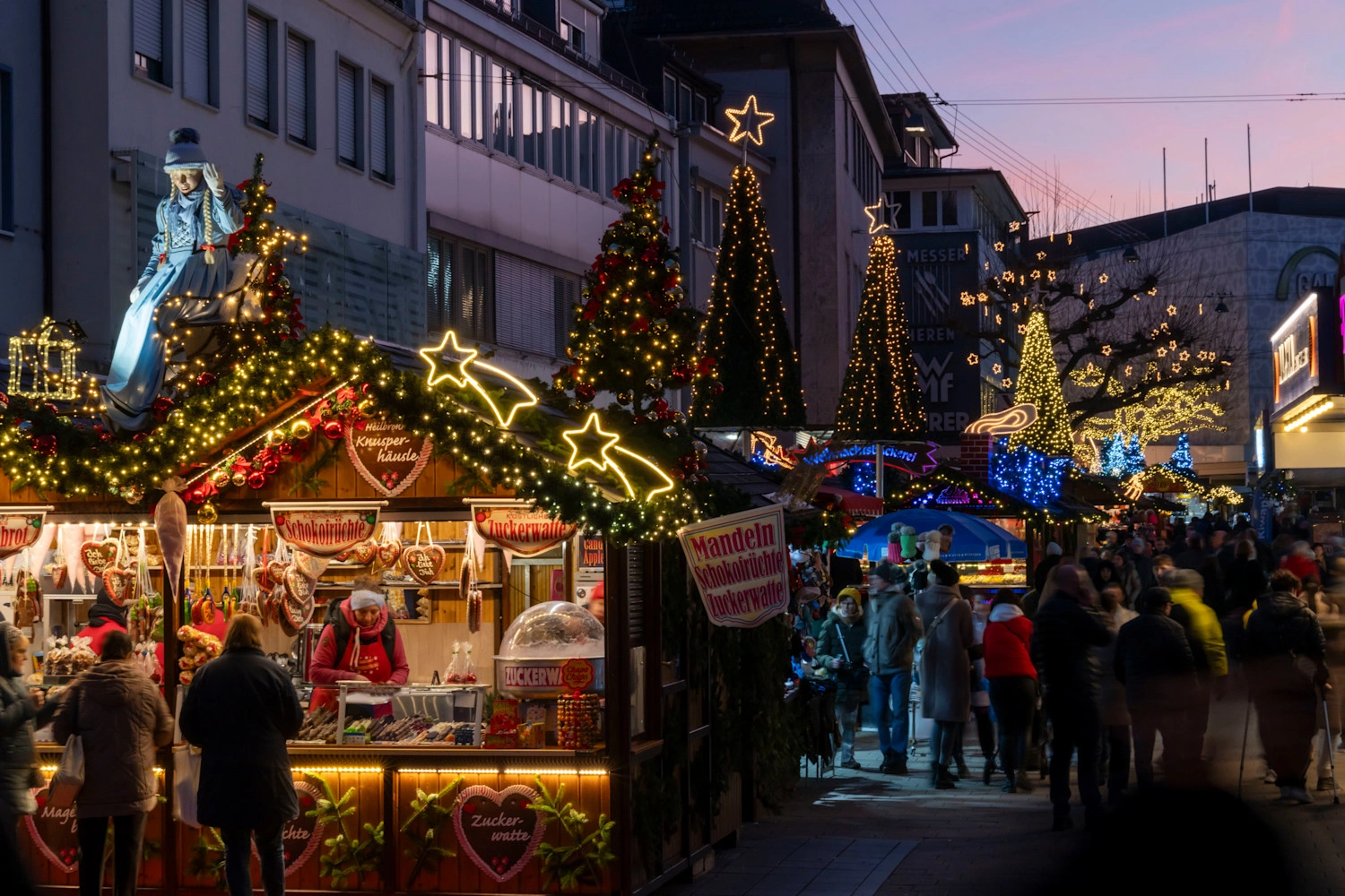 An alley of a Christmas market with illuminated stalls and decorated trees on the sides.