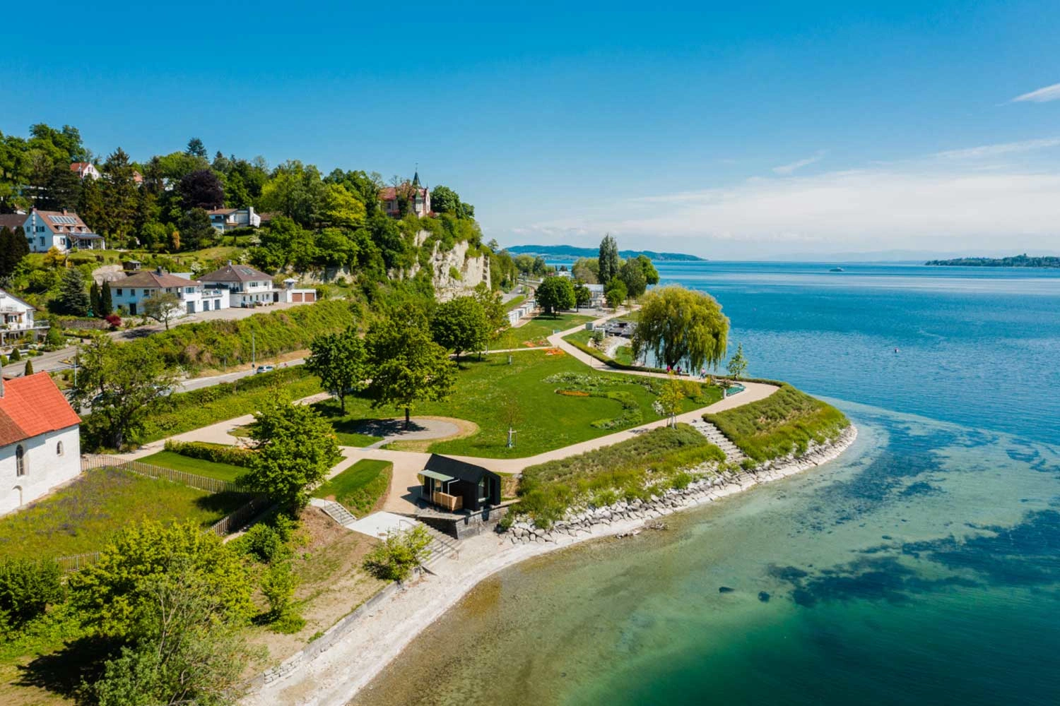 View of a park located directly on a large body of water. A village stands out behind it. The water shimmers in various shades of blue.
