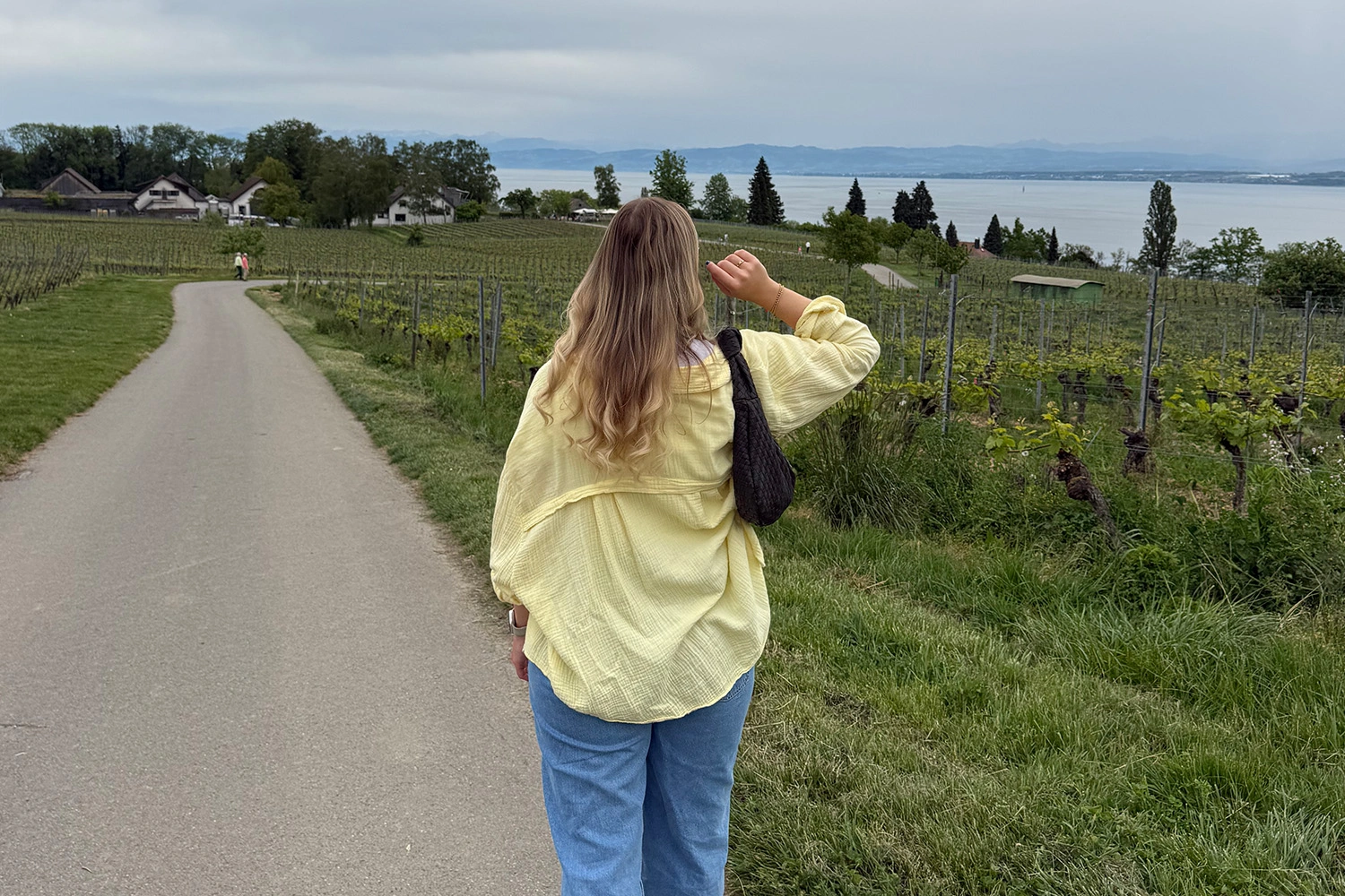 Frau spaziert durch Weinberge oberhalb des Bodensees; Panorama mit See und Alpen wirbt für Wanderwege und Naturerlebnis nahe Meersburg.
