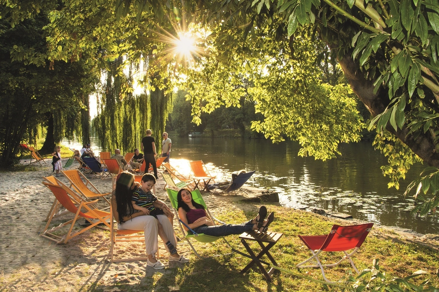Women with children sit in deckchairs on the sandy banks of a river.