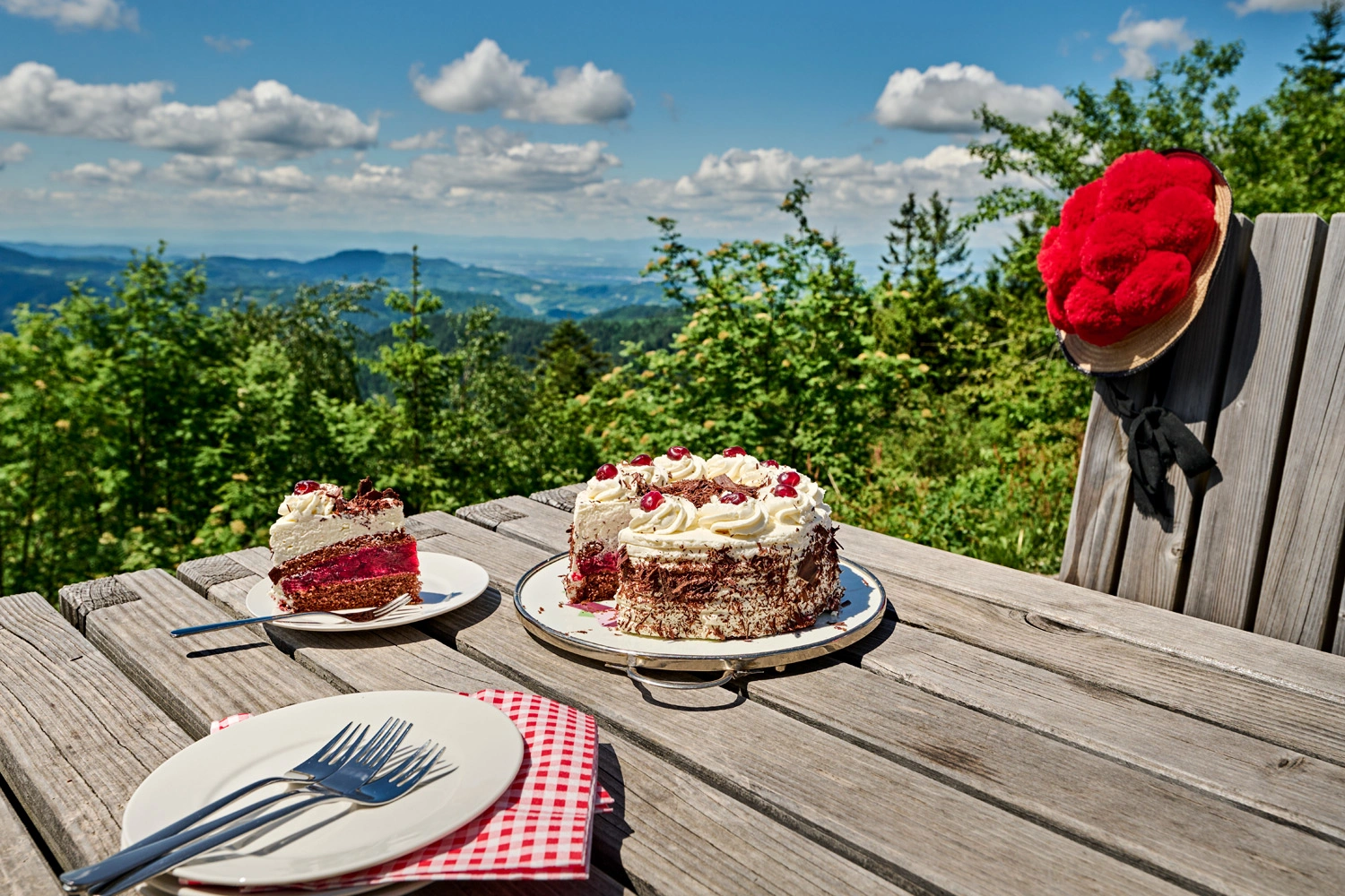 Schwarzwälder Kirschtorte mit Sahne, Kirschen und dunklen Schokoladenraspeln auf einer Platte vor klassischem Schwarzwald-Panorama