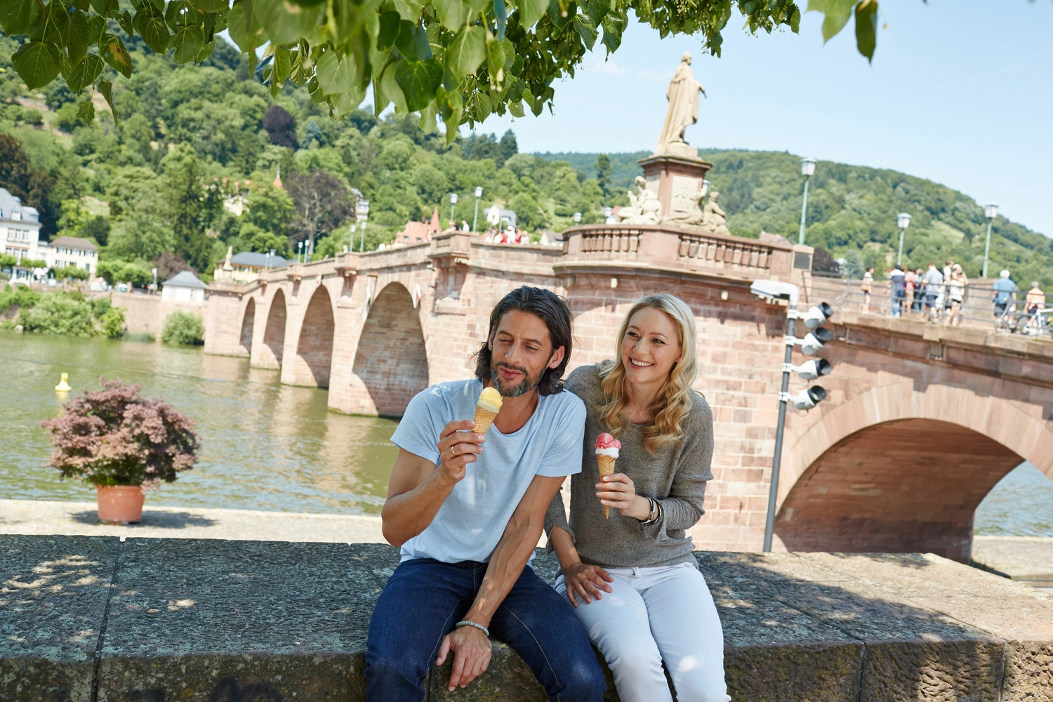 Ein Paar sitzt am Ufer eines Flusses in einer Stadt und isst Eis. Hinter ihnen befindet sich eine große Brücke über den Fluss.