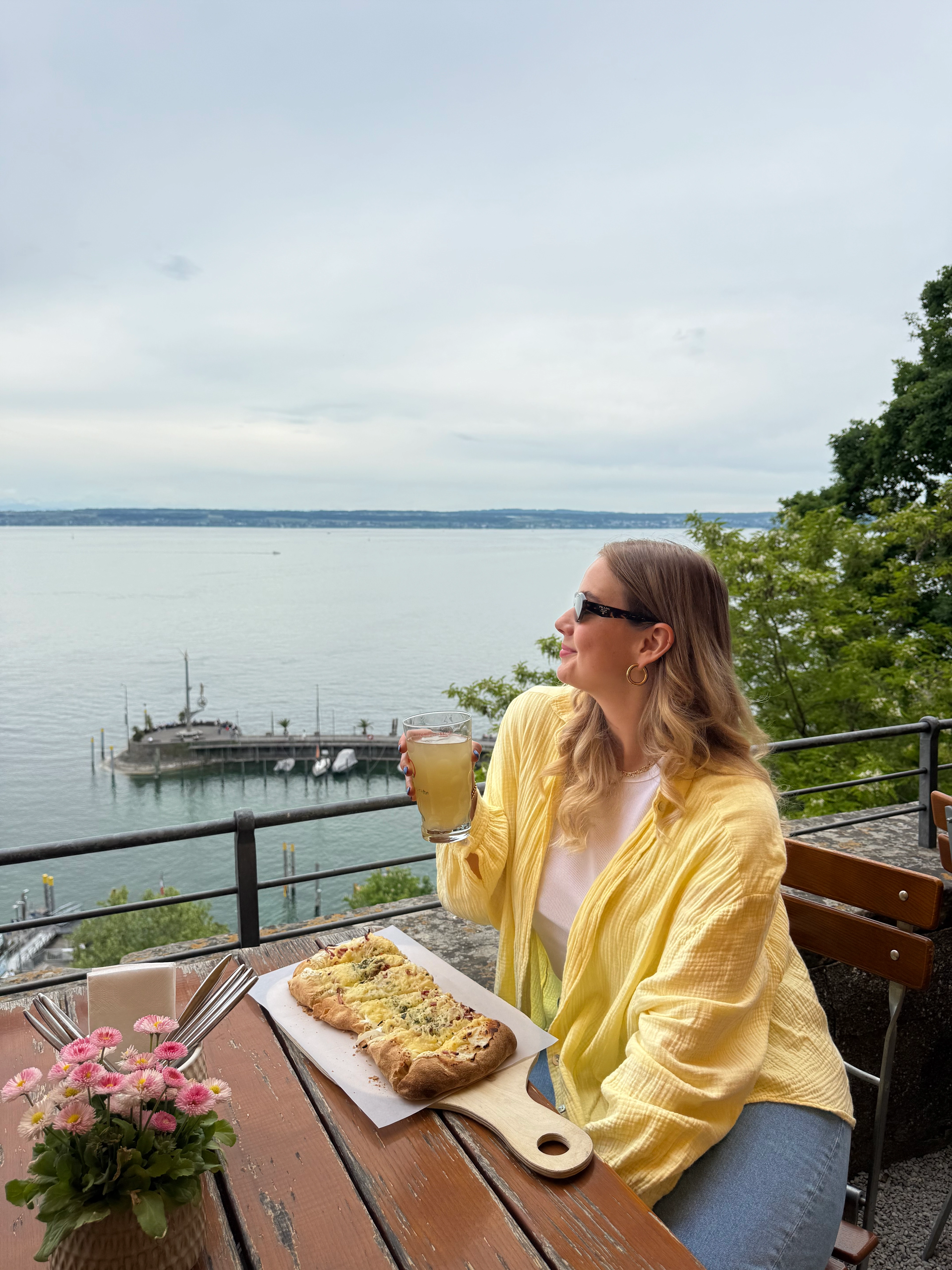 Frau auf Restaurantterrasse über dem Bodensee genießt Flammkuchen und Getränk; Blick auf See und Hafen bewirbt Kulinarik und Aussicht in Meersburg.