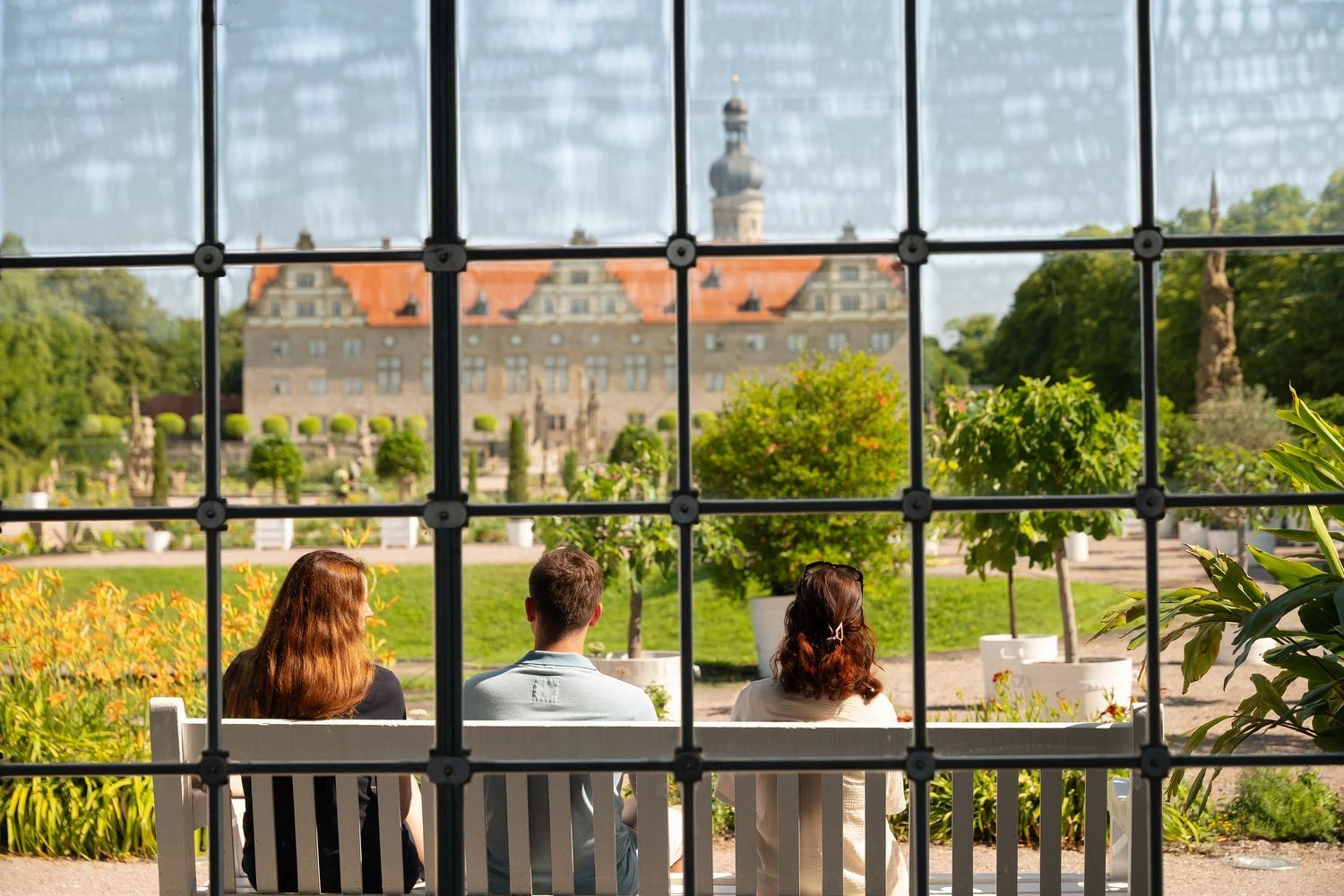 Drei Personen sitzen auf einer Bank in der Orangerie und blicken auf den barocken Schlossgarten von Weikersheim mit dem Schloss im Hintergrund.