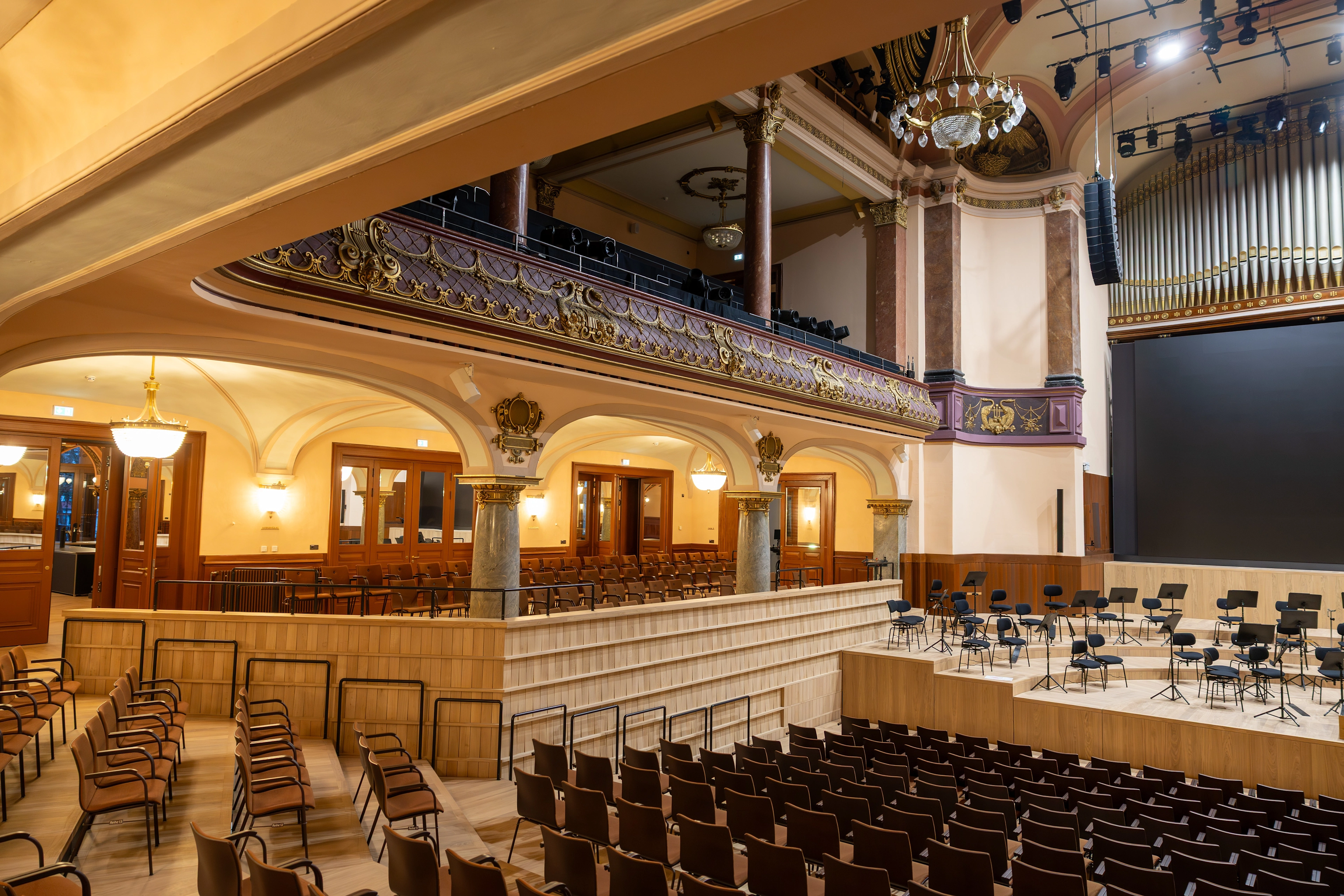 Festlich beleuchteter, historischer Konzertsaal der Stadthalle Heidelberg mit verzierten Emporen, Säulen und Orgel; davor moderne Bühne mit Orchesterstühlen und Notenpulten