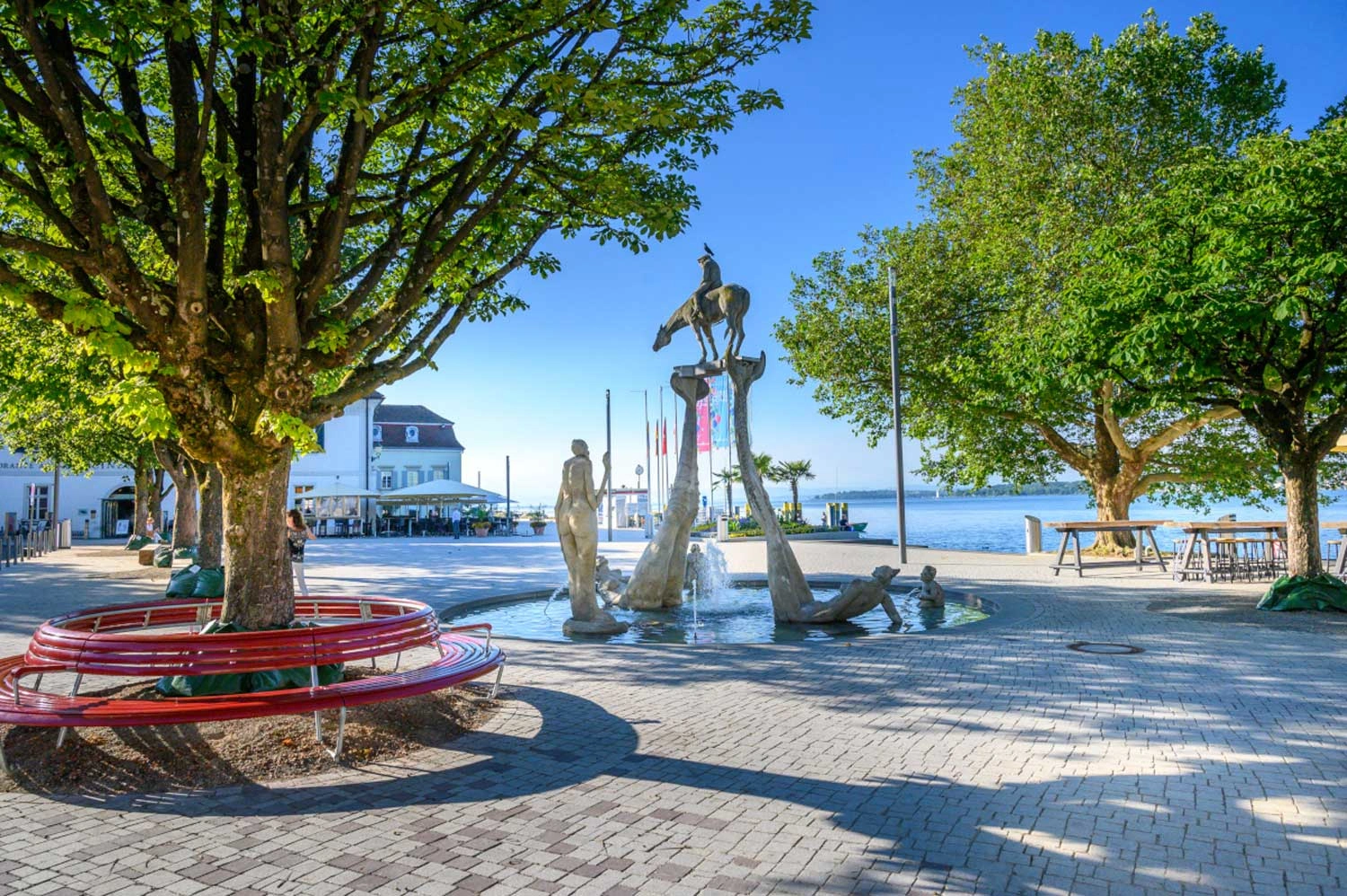 Planted square with trees. Seating areas are built around the trees. In the centre is a fountain with sculptures. There is a large lake in the background.