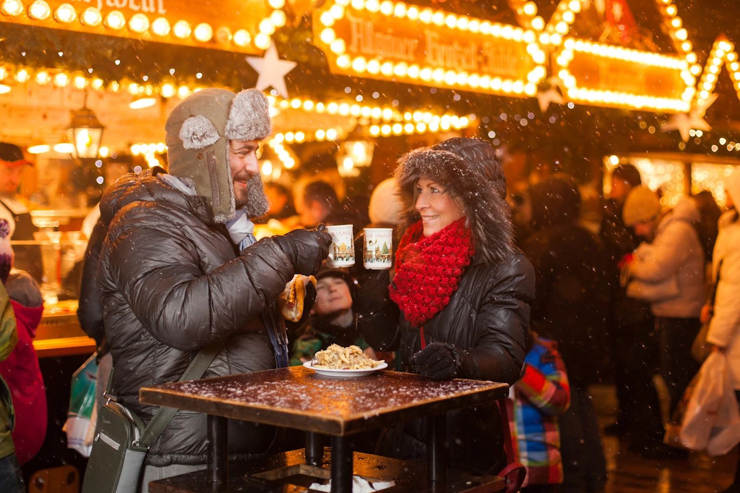 Two people in thick winter clothing stand at a Christmas market and toast with mulled wine.