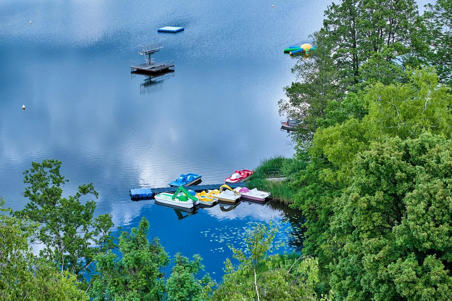 Blick auf den Obersee in Kißlegg mit Tretbooten, Badeplattform und Uferbäumen.