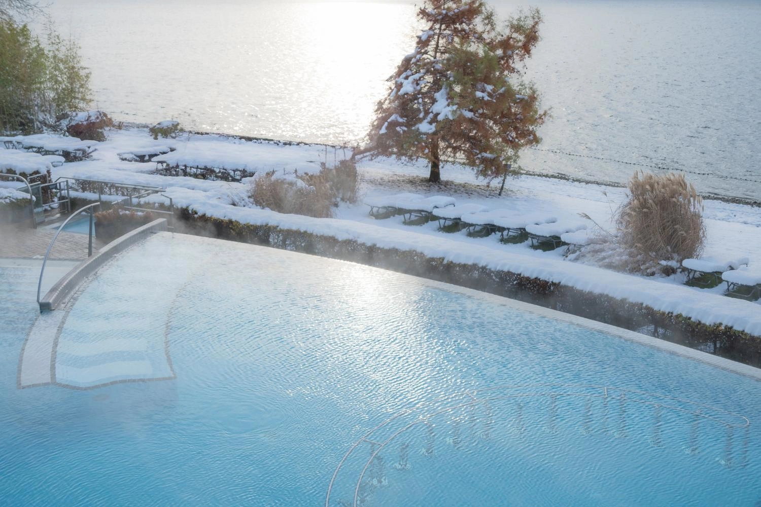 Außenpool mit dampfendem Wasser am verschneiten Seeufer, umgeben von Winterlandschaft und klarem Wasserblick.
