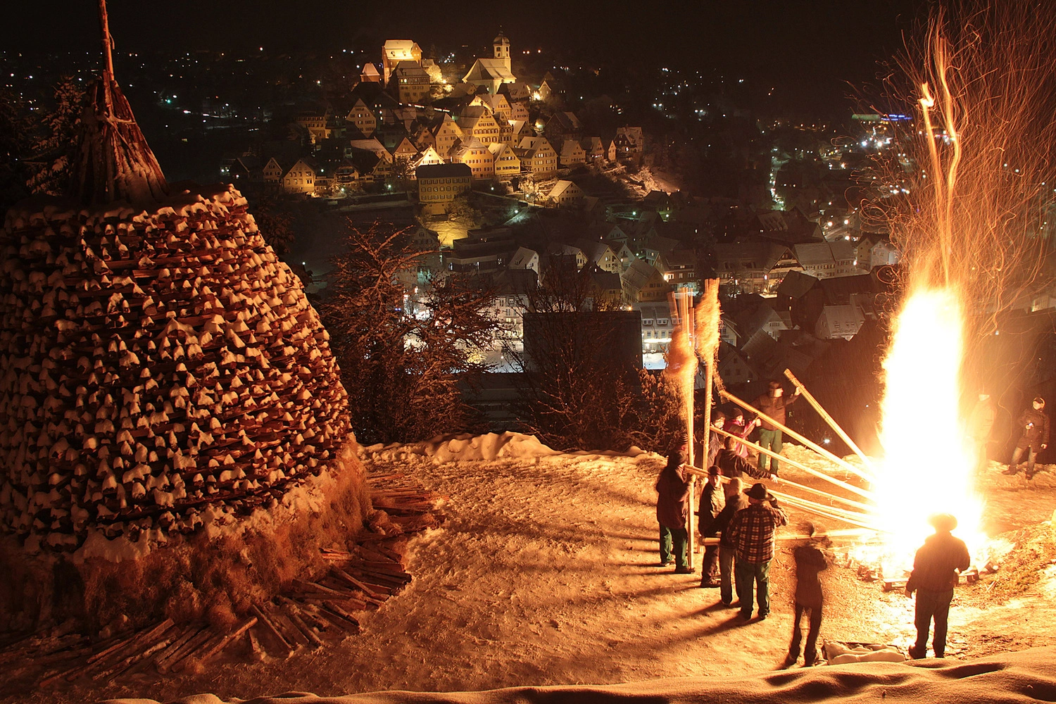 Menschen entzünden ein Weihnachtsfeuer in der Dunkelheit. Im Hintergrund eine beleuchtete Stadt.