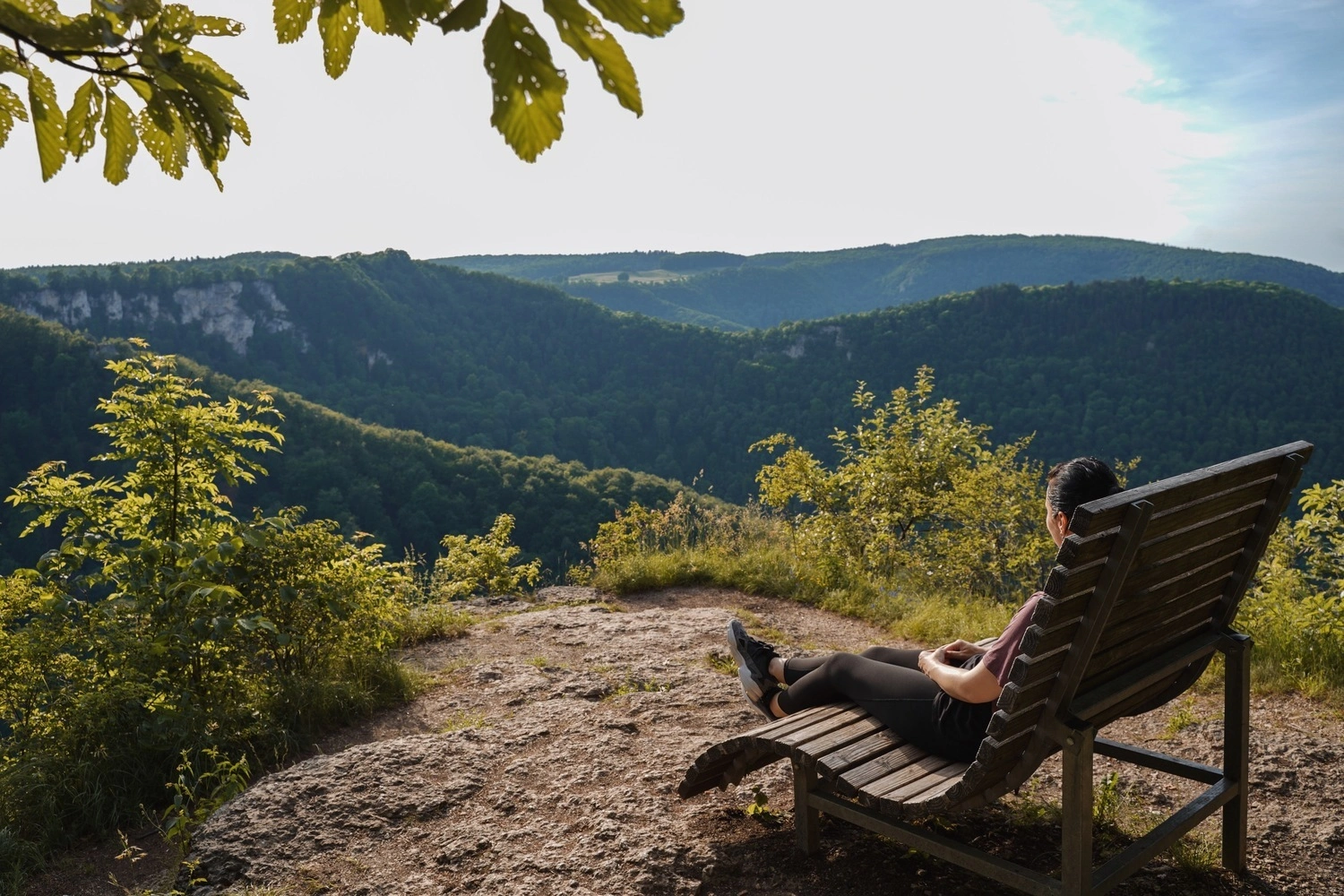 A woman is sitting on a wooden deckchair at a viewpoint, enjoying the view over the rolling countryside.