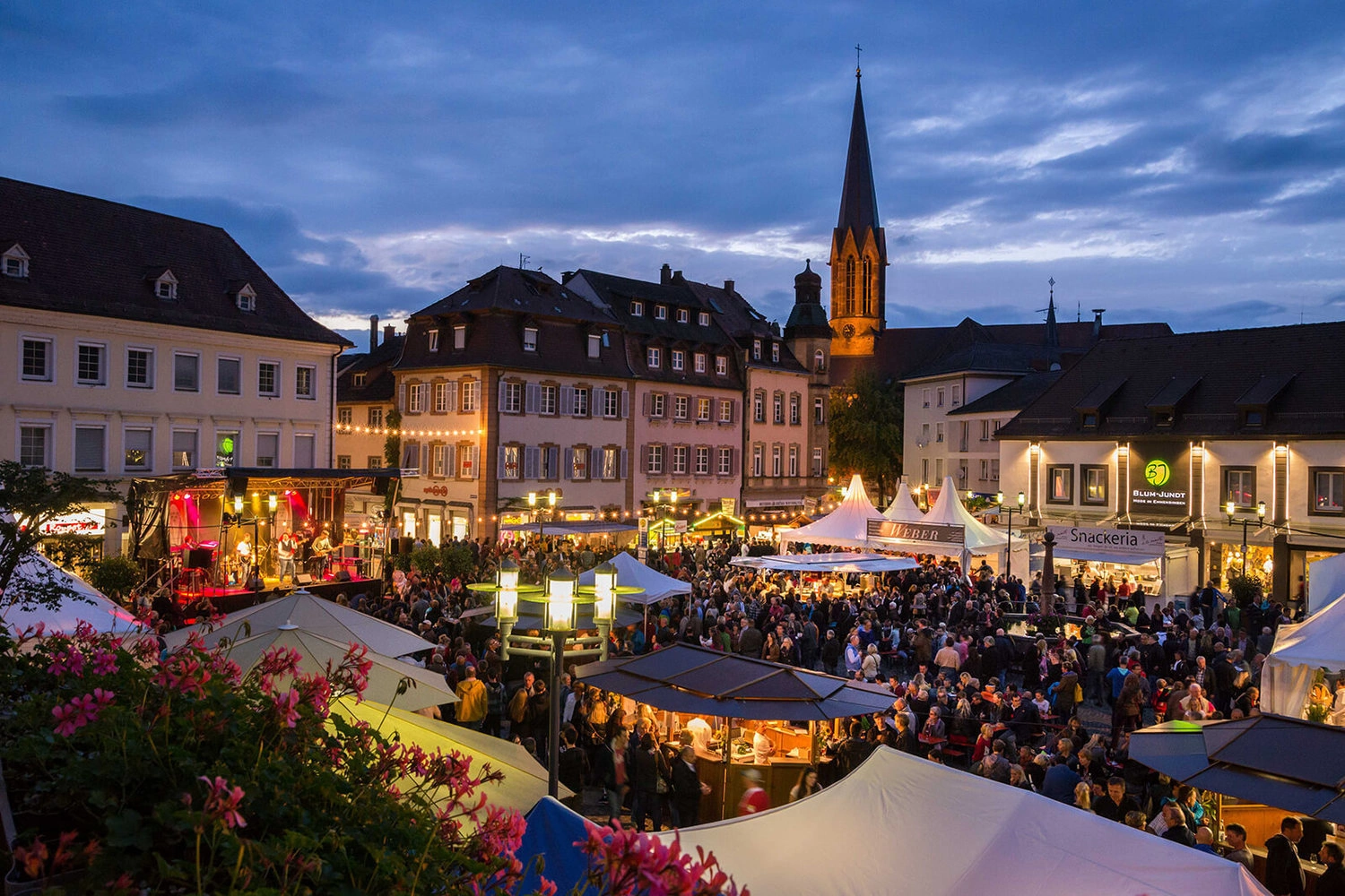 Großer, beleuchteter Platz in einer Stadt mit vielen Ständen und Menschen.