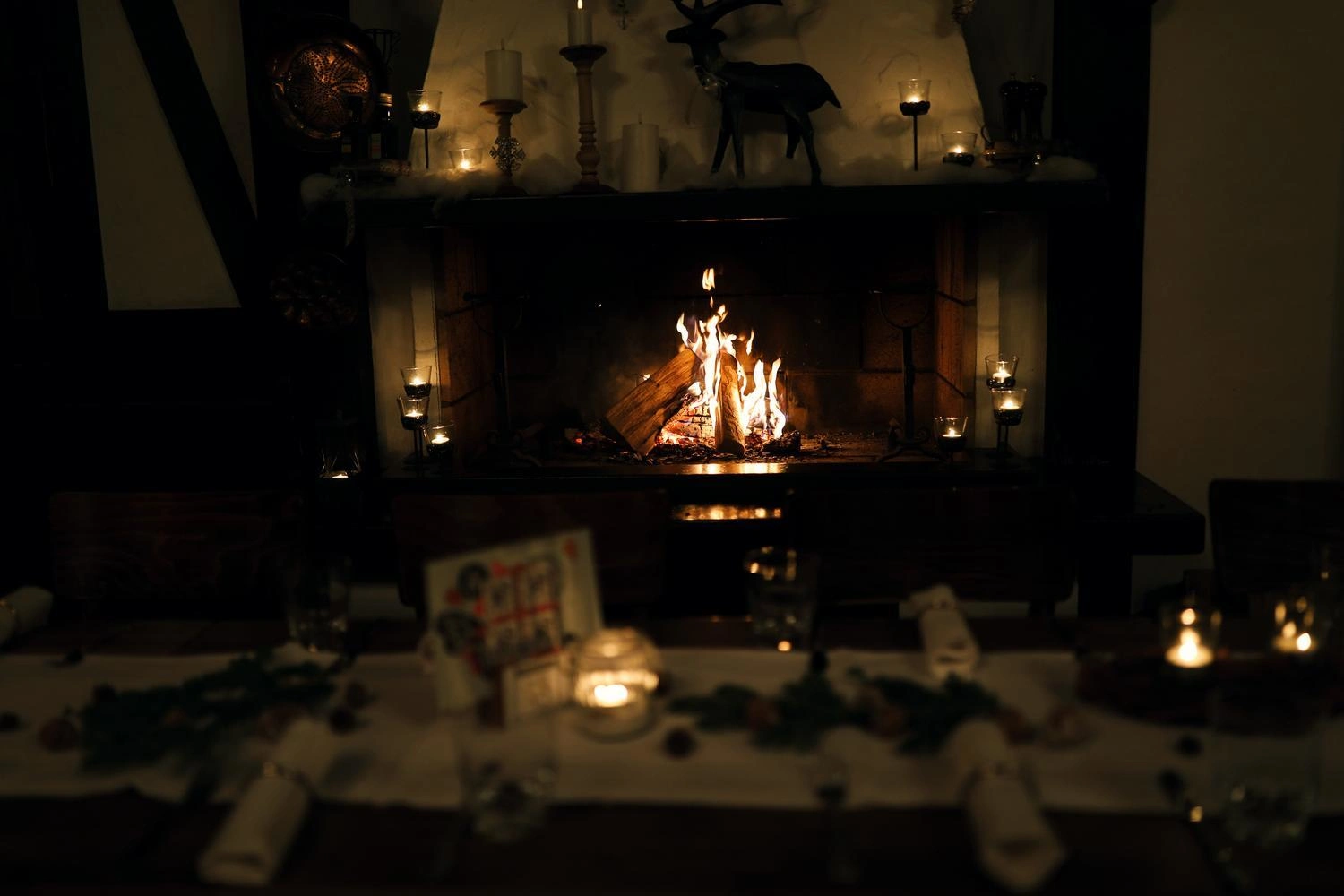 Gemütlicher Kamin mit loderndem Feuer, umgeben von Kerzen und Dekoration, festlich gedeckter Tisch im Vordergrund.