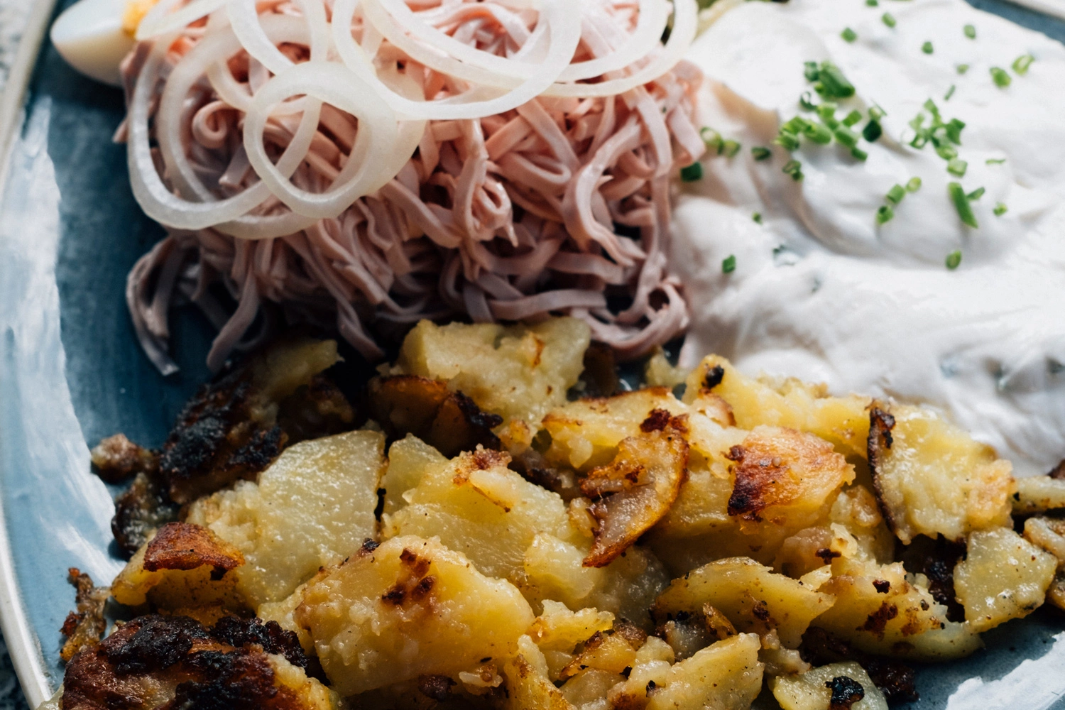Traditionelles badisches Dreierlei: knusprige Bratkartoffeln, feiner Wurstsalat mit Zwiebelringen und cremiger Quark mit Schnittlauch auf einem Teller.