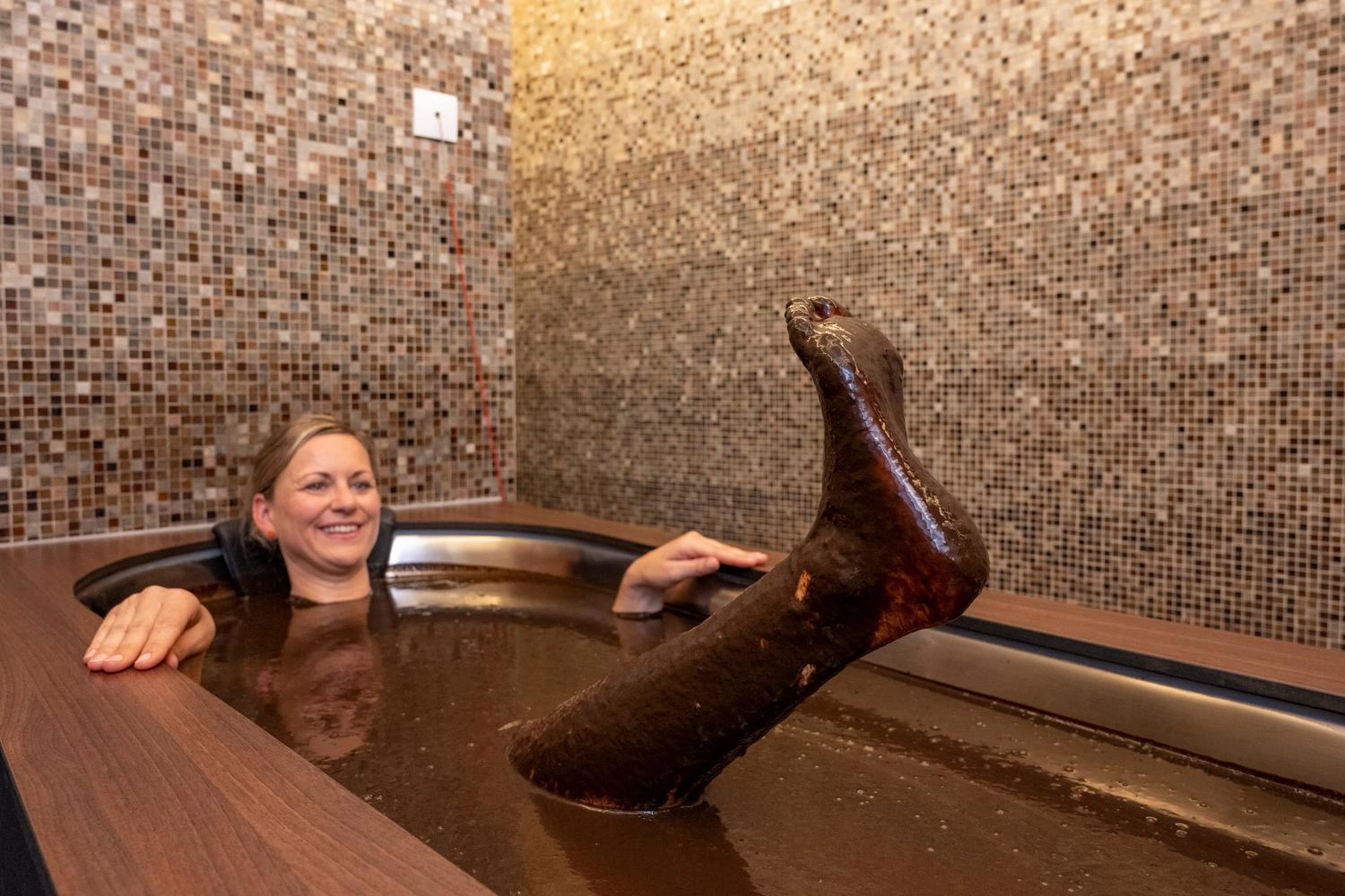 A person relaxes in a dark moor bath, one leg clearly protruding from the thick, brown liquid, surrounded by mosaic-covered walls.