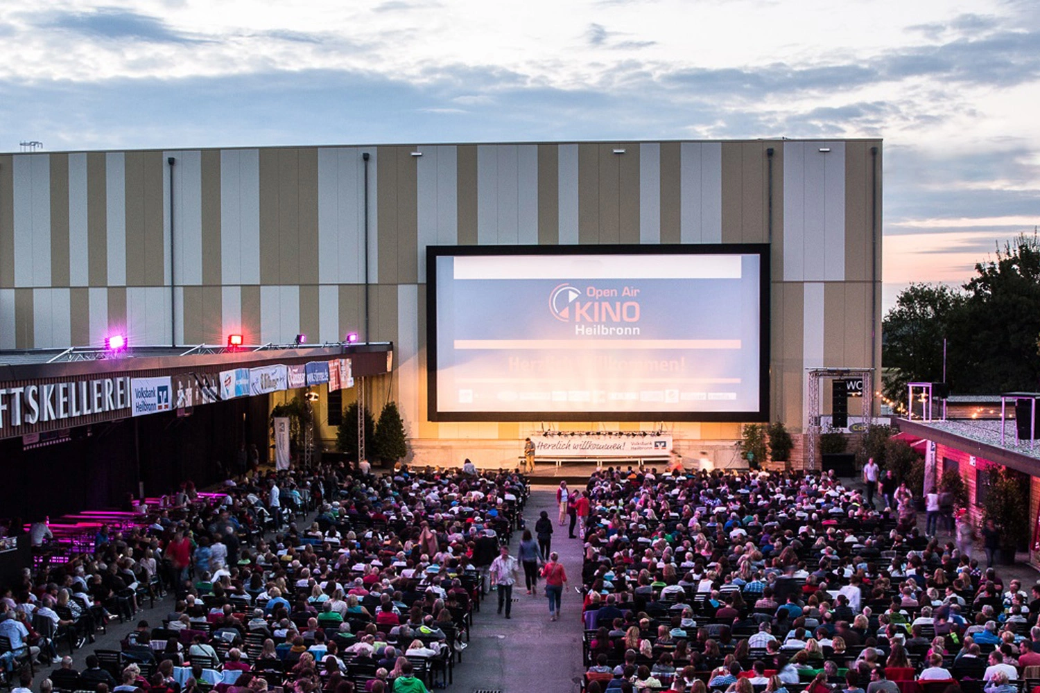 A huge screen hangs on the wall of a building. Many people sit in two rows in front of it and watch a film as dusk falls.