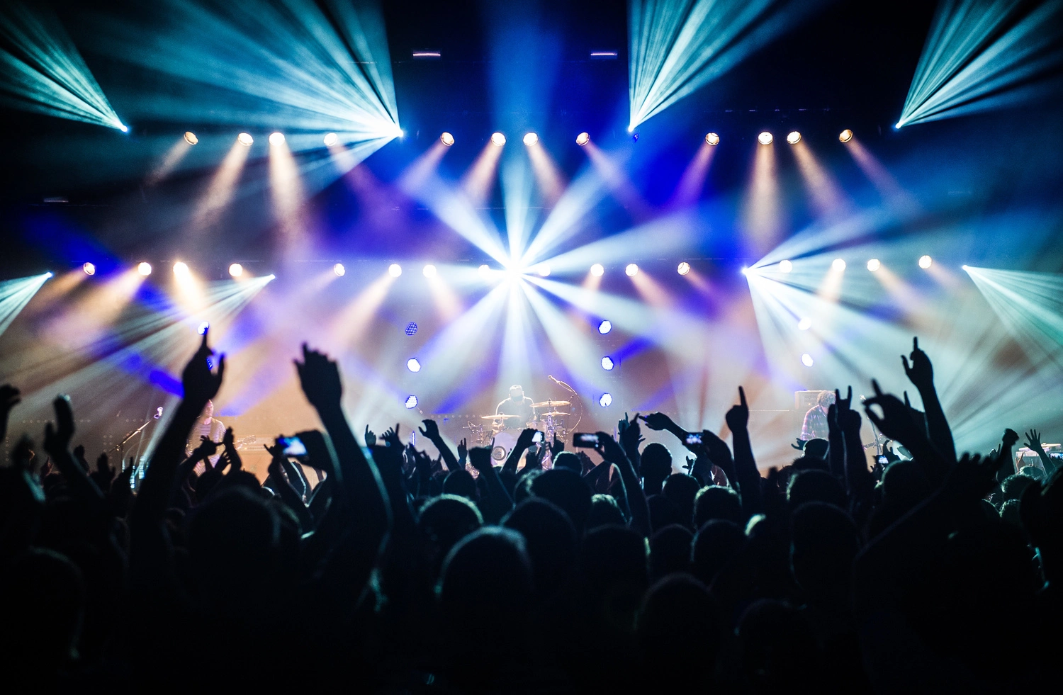 Live-Konzert mit Band auf beleuchteter Bühne, Publikum hebt Hände und Smartphones; symbolisiert Stimmung und Atmosphäre einer Musikveranstaltung auf der Eventseite.