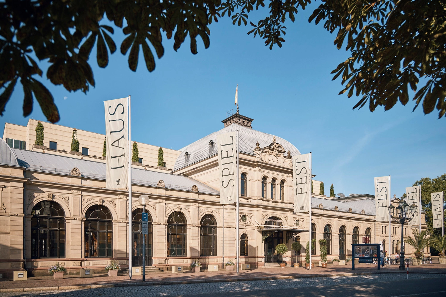 Historisches Festspielhaus mit heller Fassade, großen Rundbogenfenstern und mehreren weißen Fahnen mit der Aufschrift „FESTSPIELHAUS“ vor blauem Himmel, eingerahmt von Bäumen.
