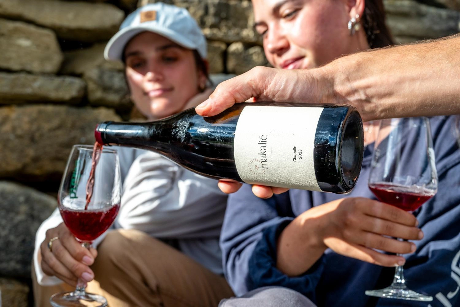 Red wine is poured from a bottle into a glass, with a stone wall and more glasses in the background.