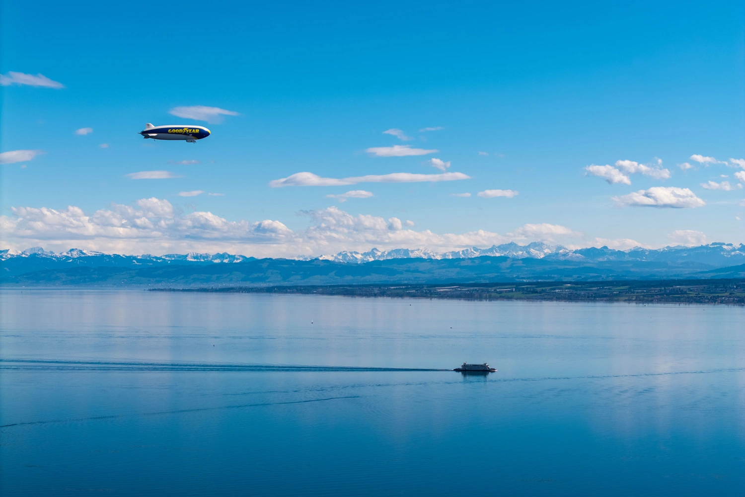 A zeppelin flies over the tranquil Lake Constance, a ship leaves its wake below, and snow-capped mountains rise in the background.