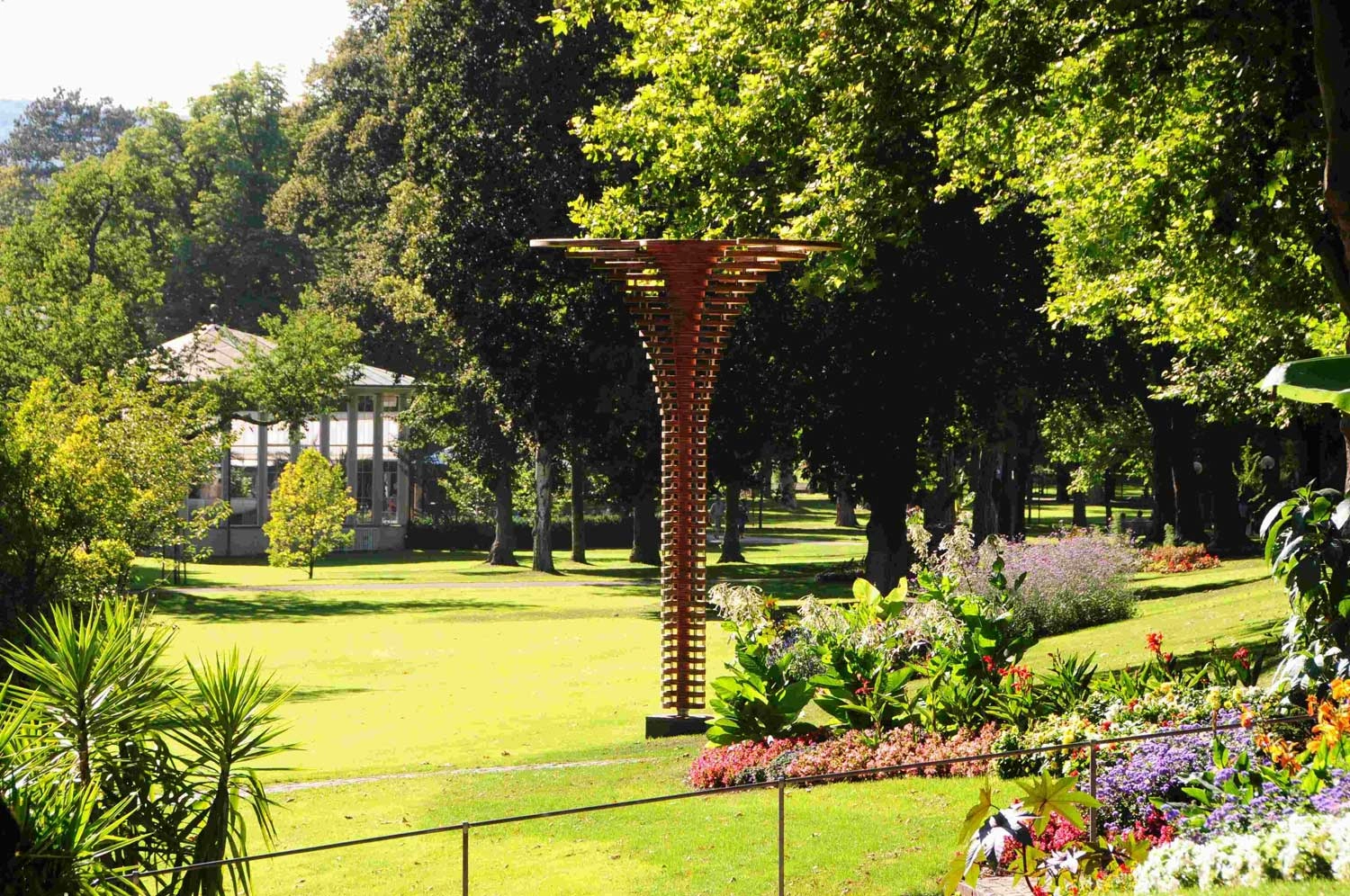 View of a meticulously landscaped park with many trees, flowerbeds and a sculpture.