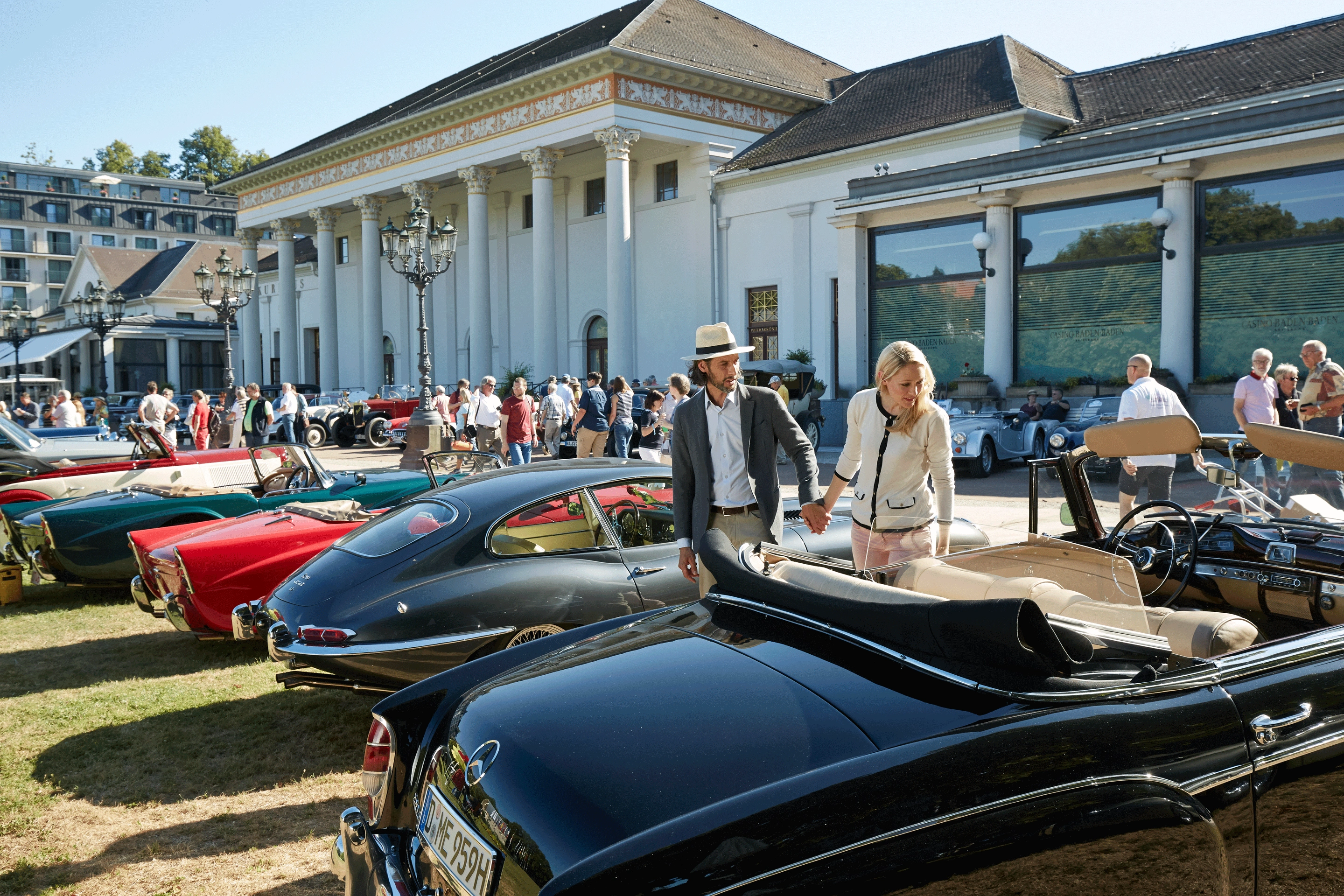  Elegantes Paar spaziert zwischen historischen Oldtimern auf der Wiese vor dem weißen Kurhaus in Baden-Baden bei Sonnenschein.