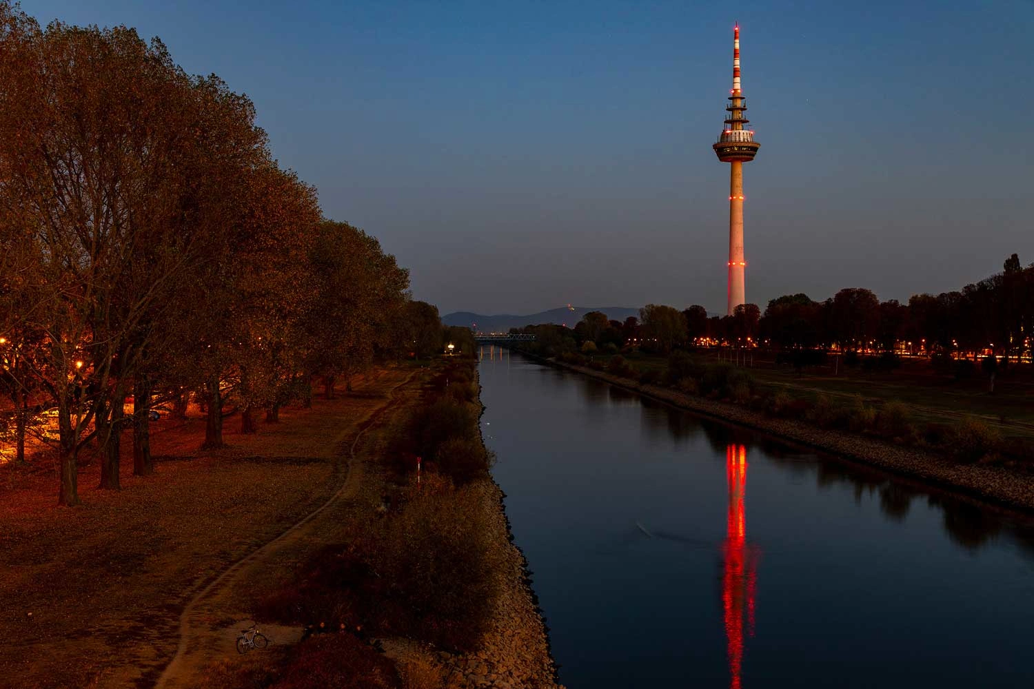 Ein Fluss mit einer Allee am Ufer bei Dämmerung, im Hintergrund ein Fernsehturm.