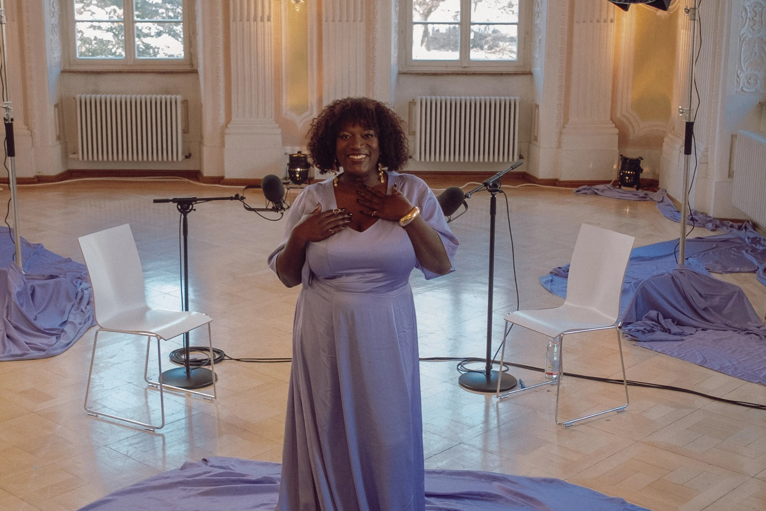 Person in elegantem, fließendem Kleid steht in einem hellen, barocken Saal zwischen zwei weißen Stühlen und Mikrofonen, umgeben von lilafarbenen Stoffbahnen auf dem Boden.