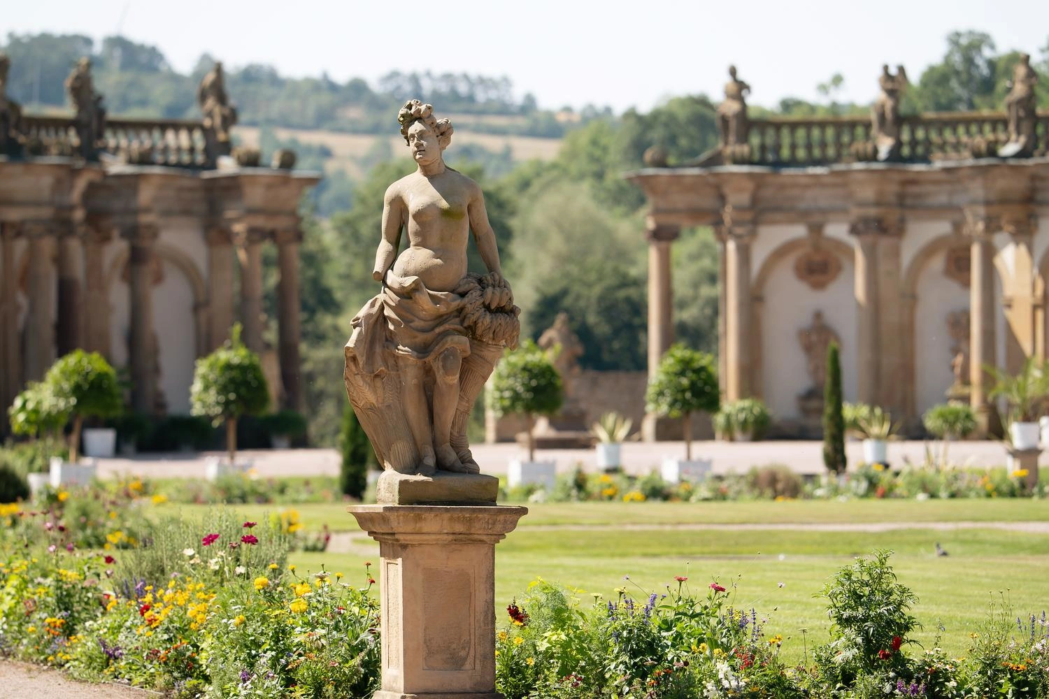 Steinskulptur in einem barocken Schlossgarten, umgeben von Blumenbeeten und Orangerie mit dekorativen Säulen im Hintergrund.